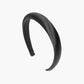 The Tori Satin Headband, made from smooth black silk, has a slightly padded surface for maximum comfort. Its minimalist and curved design allows it to fit seamlessly on the head, elegantly displayed against a white background.