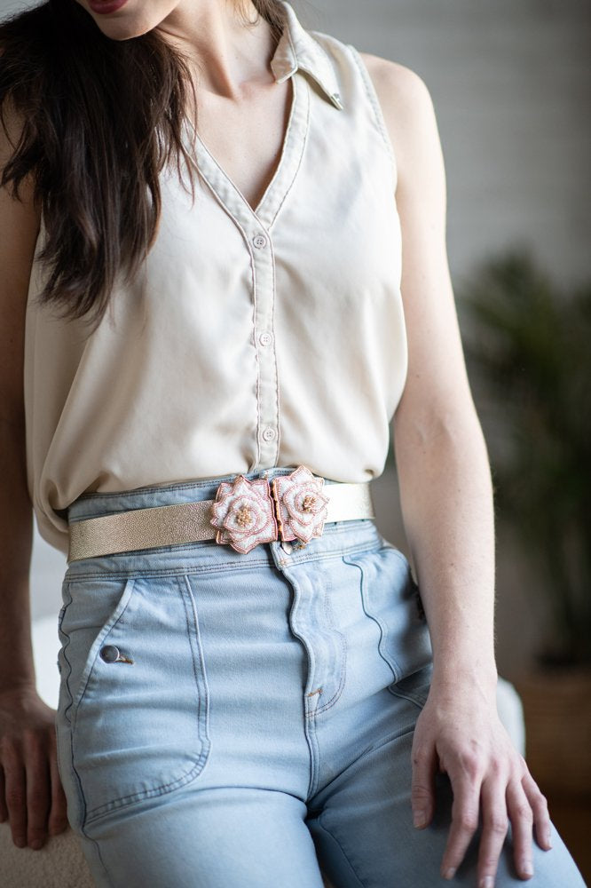 A person is wearing a sleeveless, light beige blouse tucked into high-waisted light blue jeans. They are accessorized with the Full Shade Belt, which features floral decorations and a purple tiger buckle, while a blurred plant can be seen in the background.