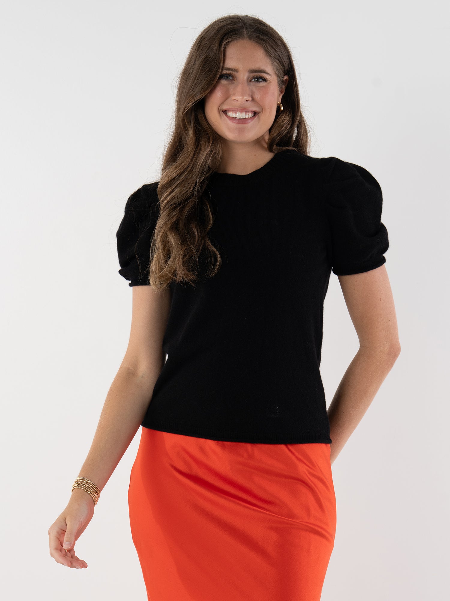 A woman with long brown hair smiles, wearing a luxurious black Crewneck Tee with puff sleeves and a bright orange skirt, standing against a plain white background.