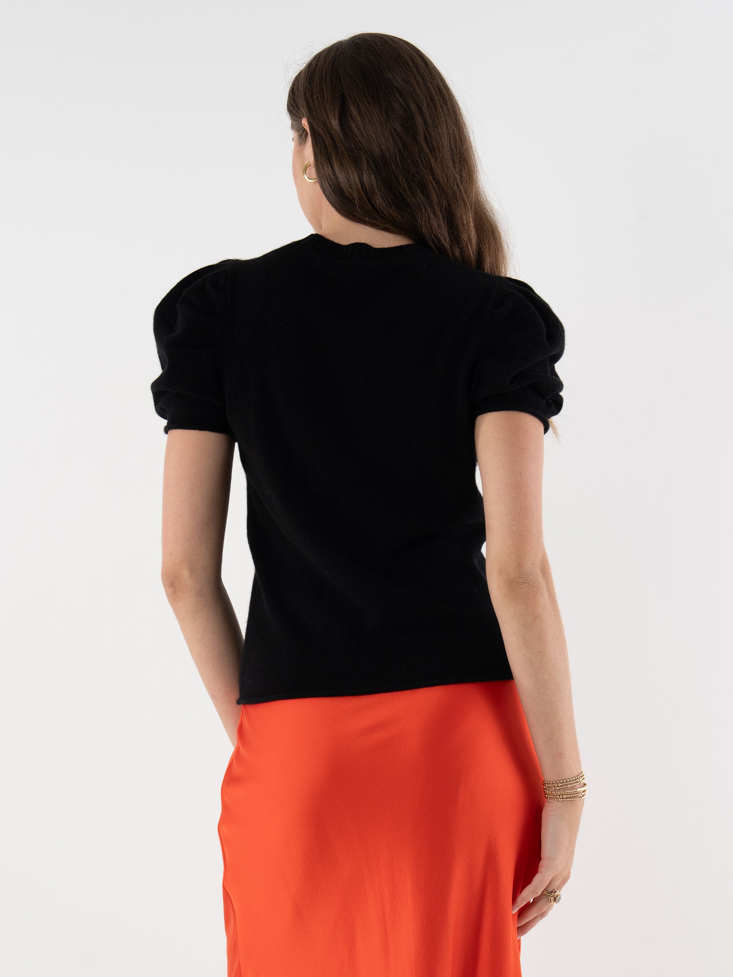 A woman with long brown hair stands with her back to the camera, wearing a black Crewneck Tee with puff sleeves and an orange skirt. She accessorizes with bracelets on her left wrist, set against a plain white background.