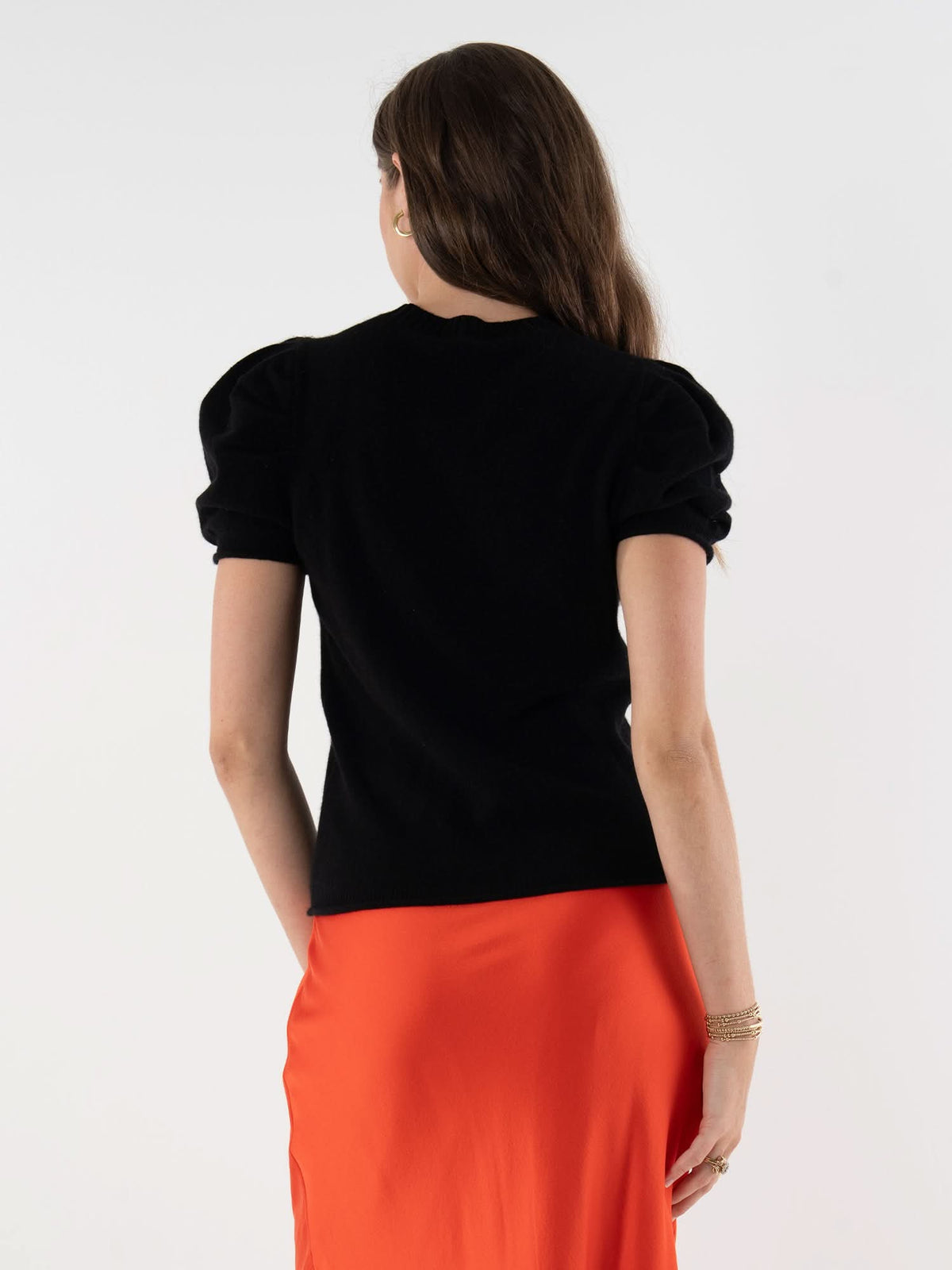 A woman with long brown hair stands with her back to the camera, wearing a black Crewneck Tee with puff sleeves and an orange skirt. She accessorizes with bracelets on her left wrist, set against a plain white background.
