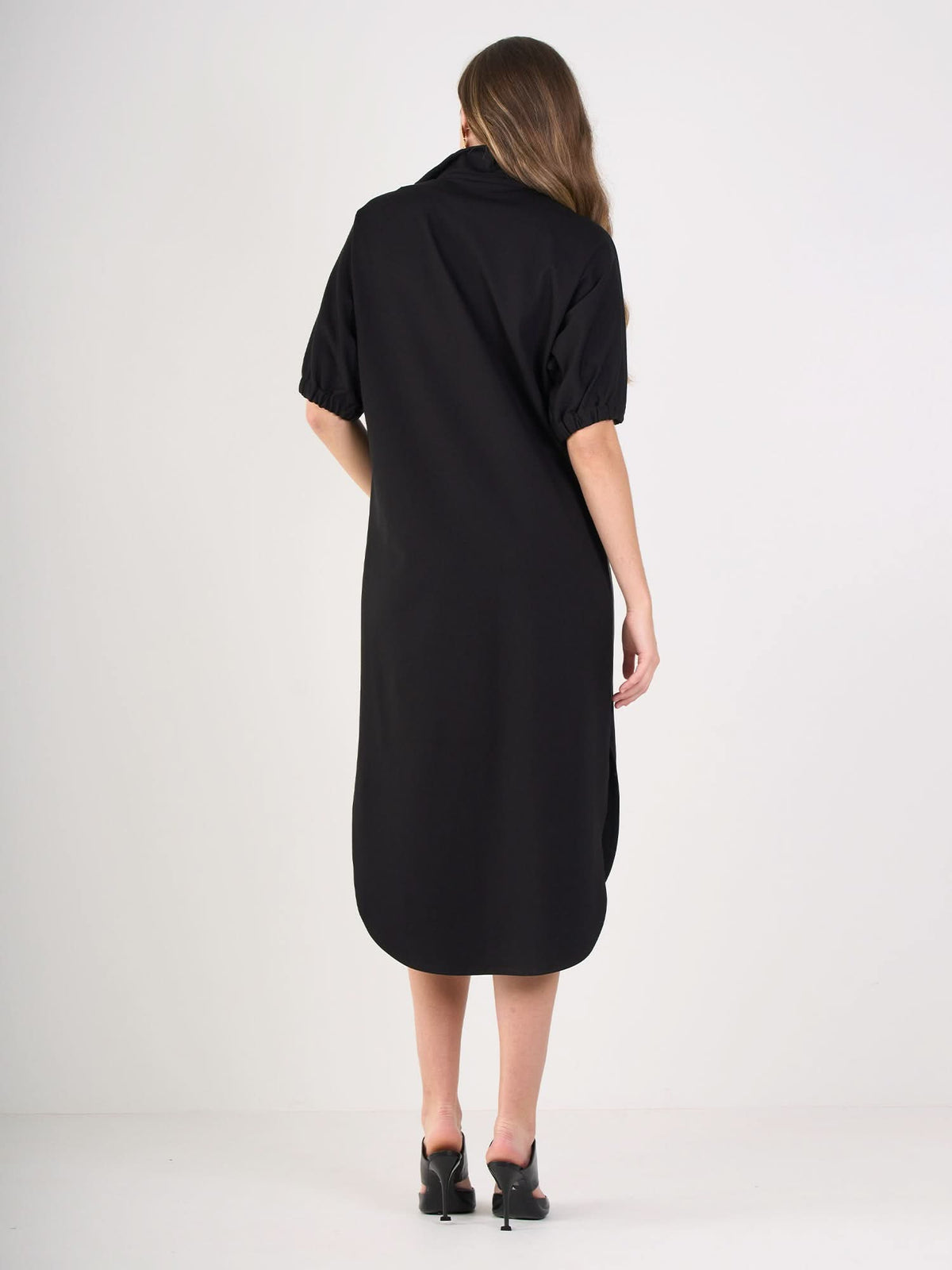 A woman with long hair models the black, loose-fitting knee-length Poppy Caftan and heeled shoes, standing with her back to the camera against a plain white background.