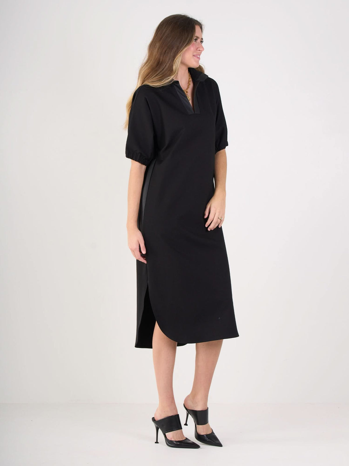A woman stands sideways, smiling, in the Poppy Caftan—a loose-fitting, knee-length black dress with short sleeves and a relaxed silhouette. She completes the look with black heeled shoes and has long, wavy hair.