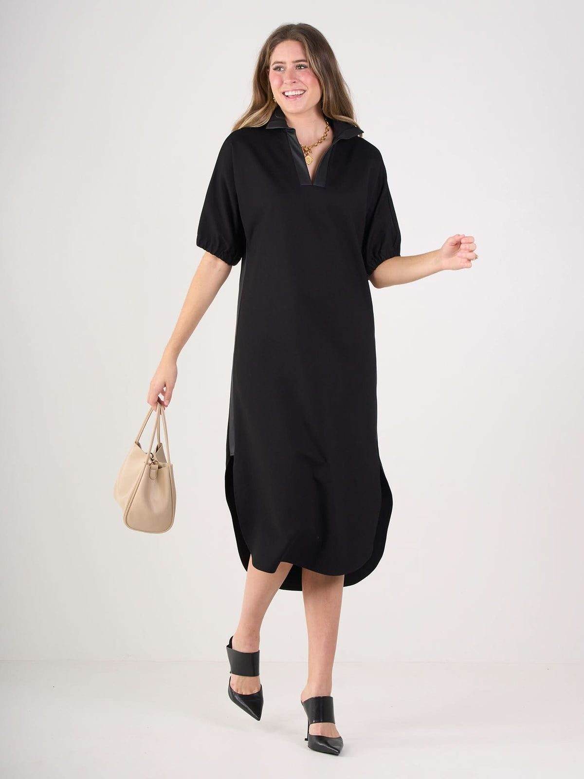 A woman wears the Poppy Caftan—a relaxed black midi dress with short sleeves and a collar. She smiles, holding a beige handbag and wearing black ankle-strap shoes. The background is plain white.