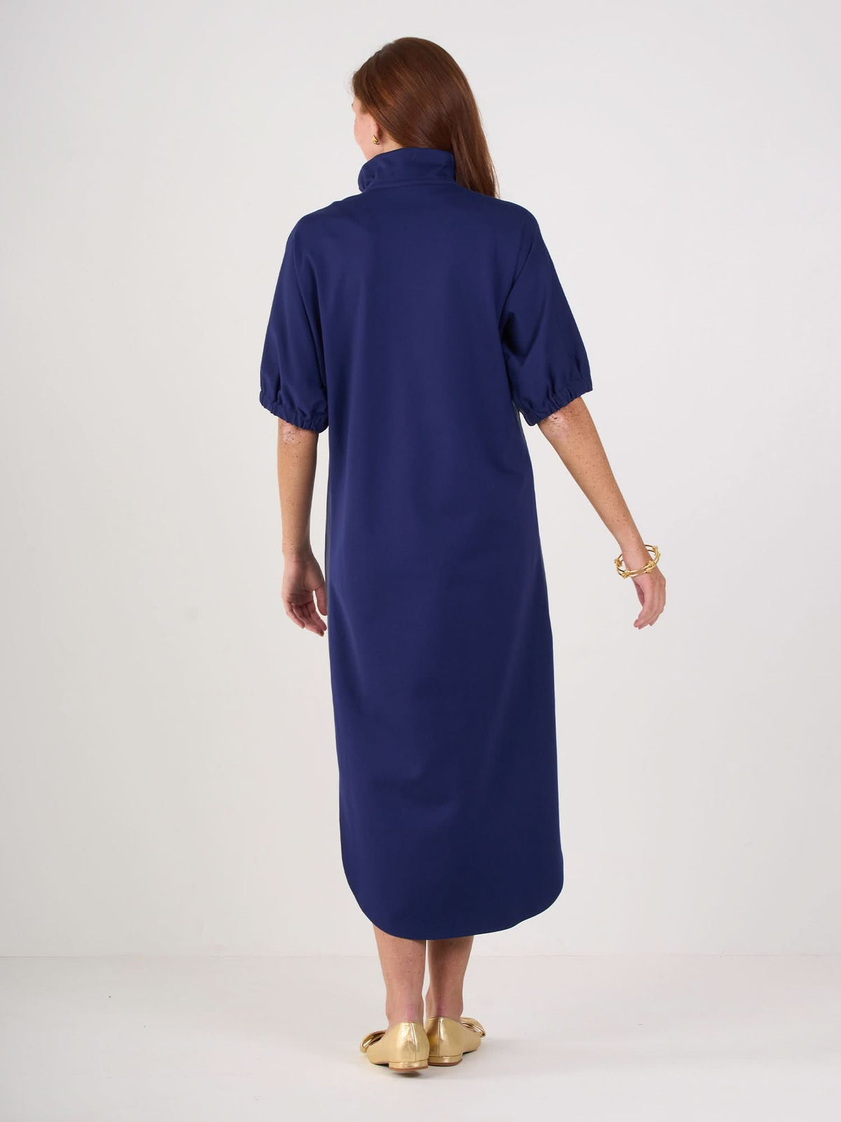 A woman with long brown hair stands facing away, wearing the Poppy Caftan—a relaxed, navy midi dress with half sleeves and a high collar—paired with a gold bracelet and gold flats, against a plain white background.