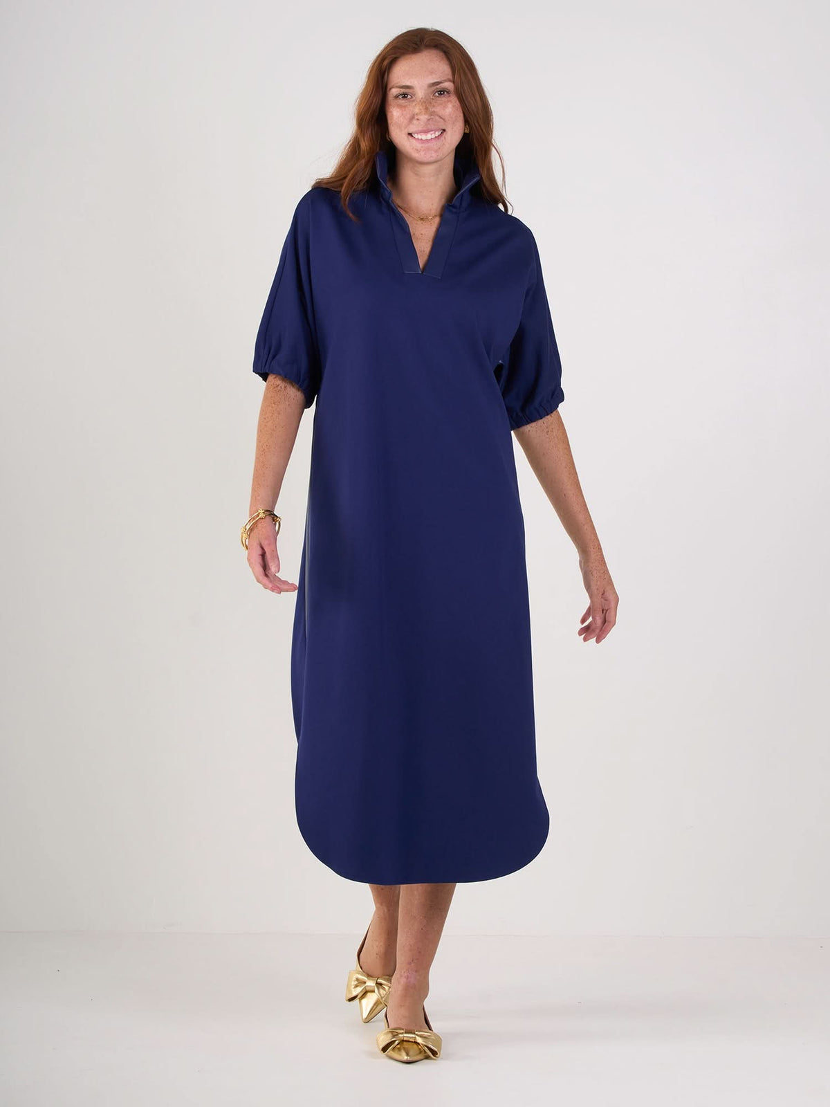 A woman with long auburn hair smiles and walks forward in the Poppy Caftan, a relaxed navy ponte style with elbow-length sleeves, paired with gold pointed-toe flats with bows, against a plain white background.