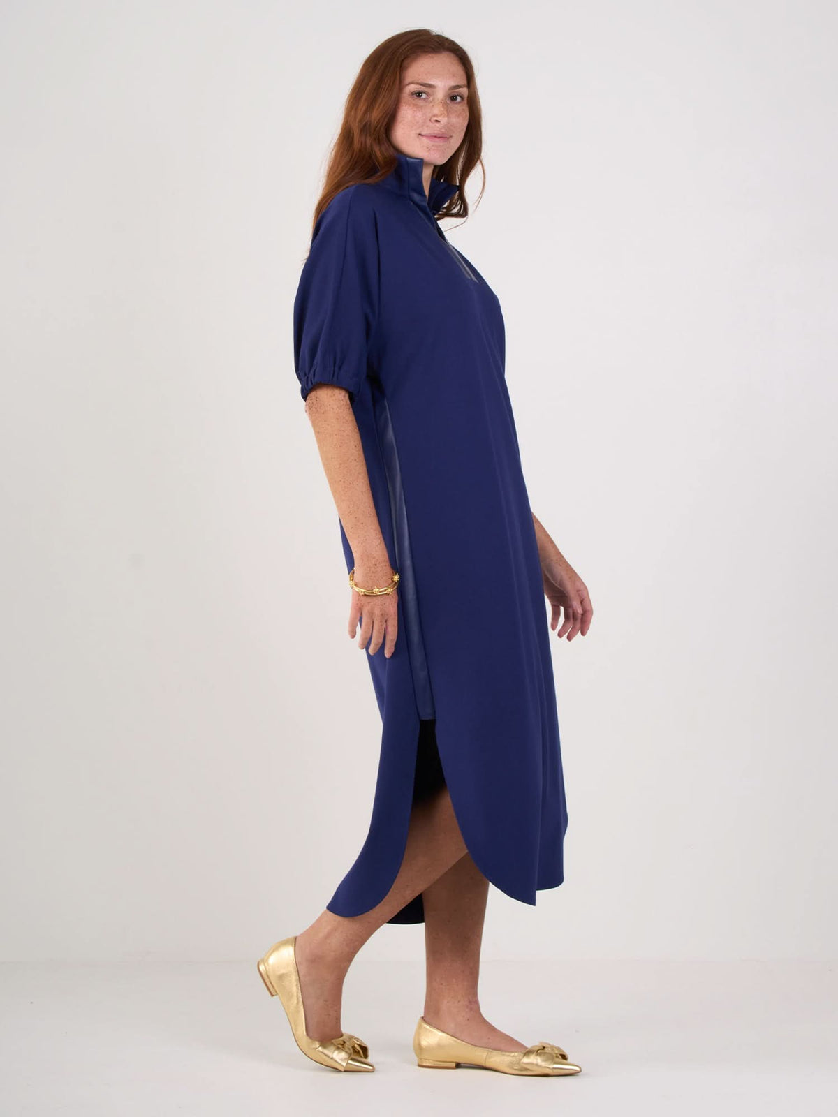 A woman with long red hair models the Poppy Caftan, a loose-fitting, knee-length royal blue dress with short sleeves and a relaxed silhouette. She stands sideways in gold flats against a plain white background.