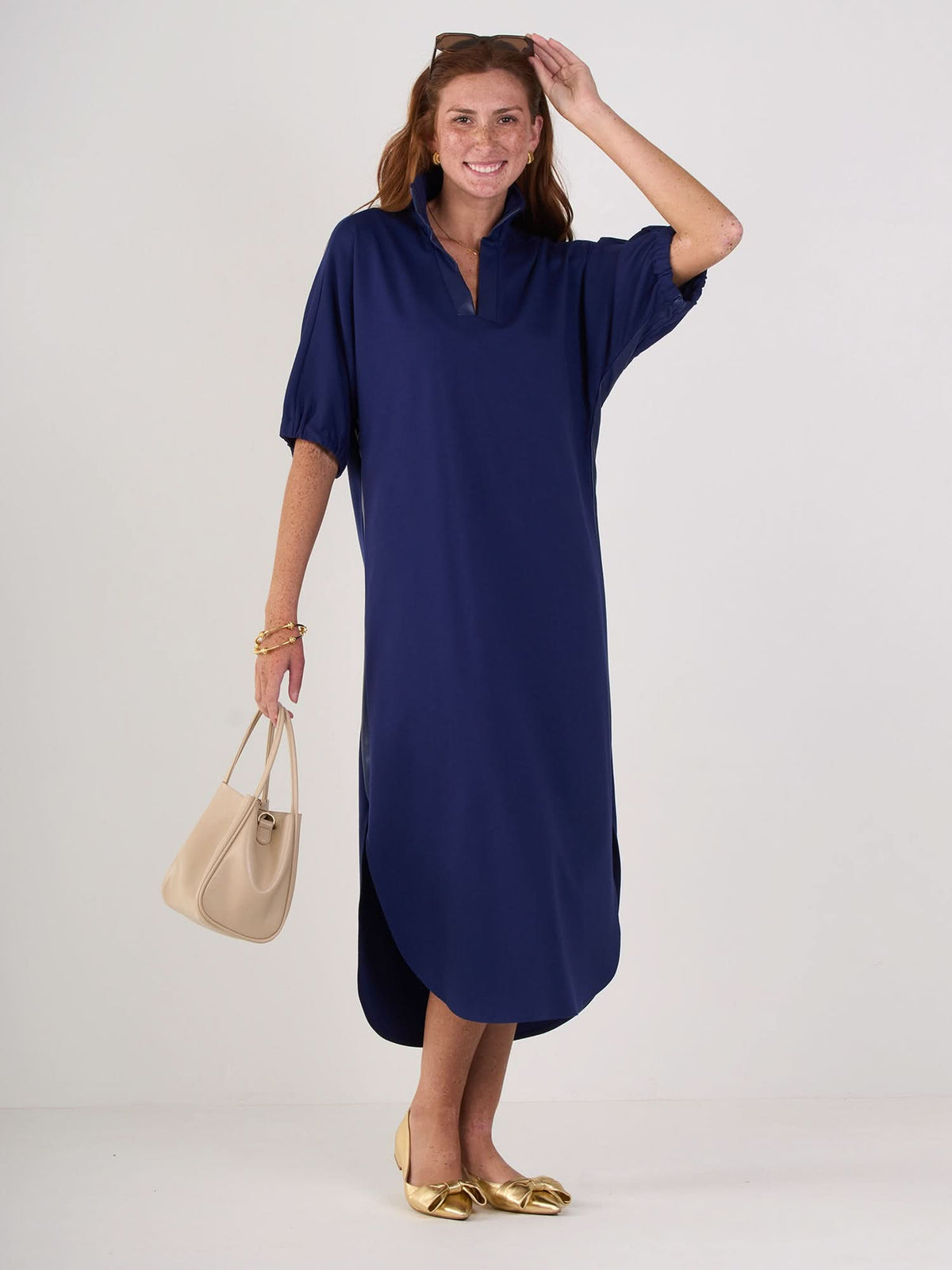 A woman stands smiling against a plain background, wearing the relaxed silhouette navy Poppy Caftan with short sleeves. She holds a beige handbag with vegan leather trim, wears gold flats, and her hair is pulled back with a headband.