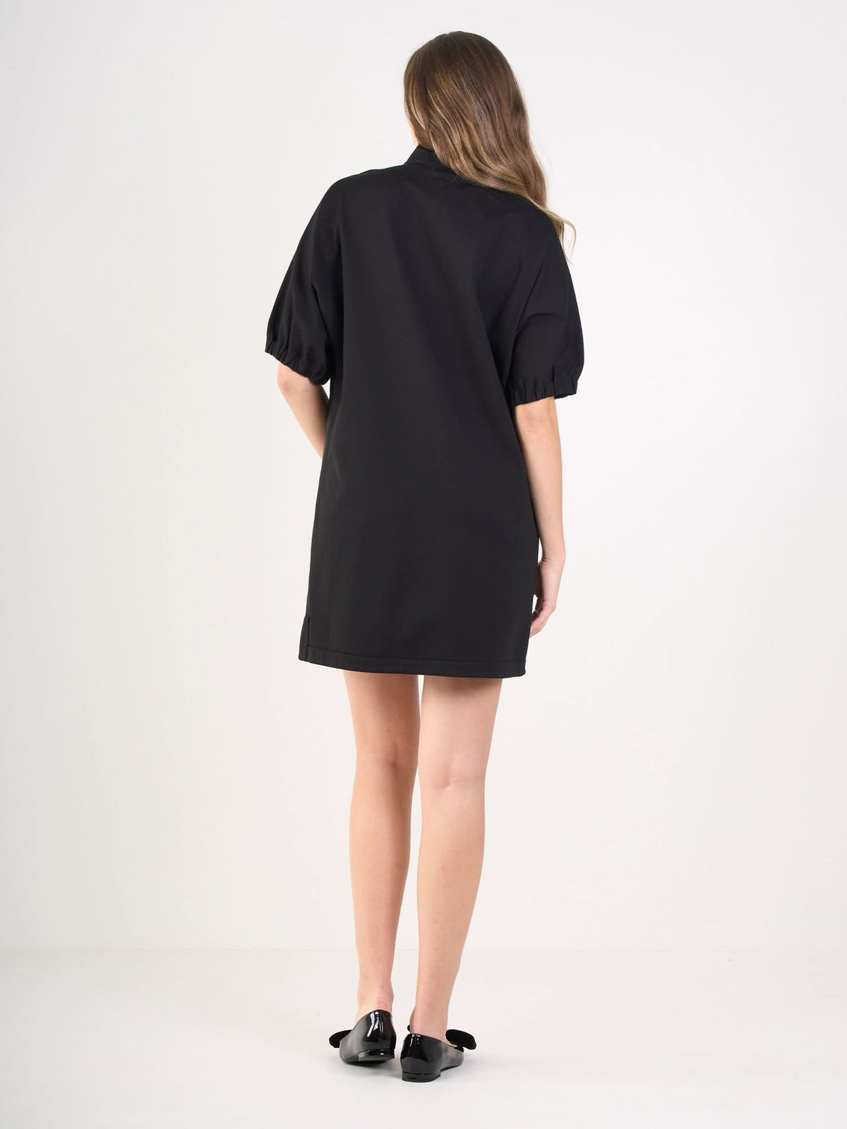 A woman with long, wavy hair faces away, wearing the Poppy Dress—a short black dress with puffed sleeves—and black flats, standing against a plain white background.