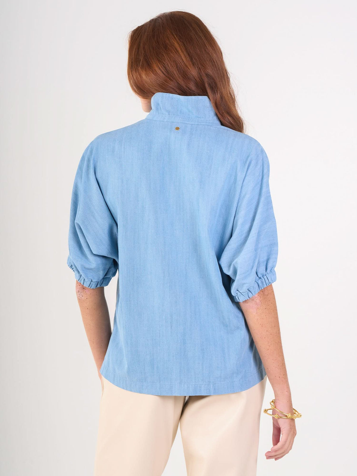 A person with long red hair is shown from the back, wearing the light blue Poppy Top with a high collar and elbow-length puff sleeves, paired with beige pants against a plain white background.