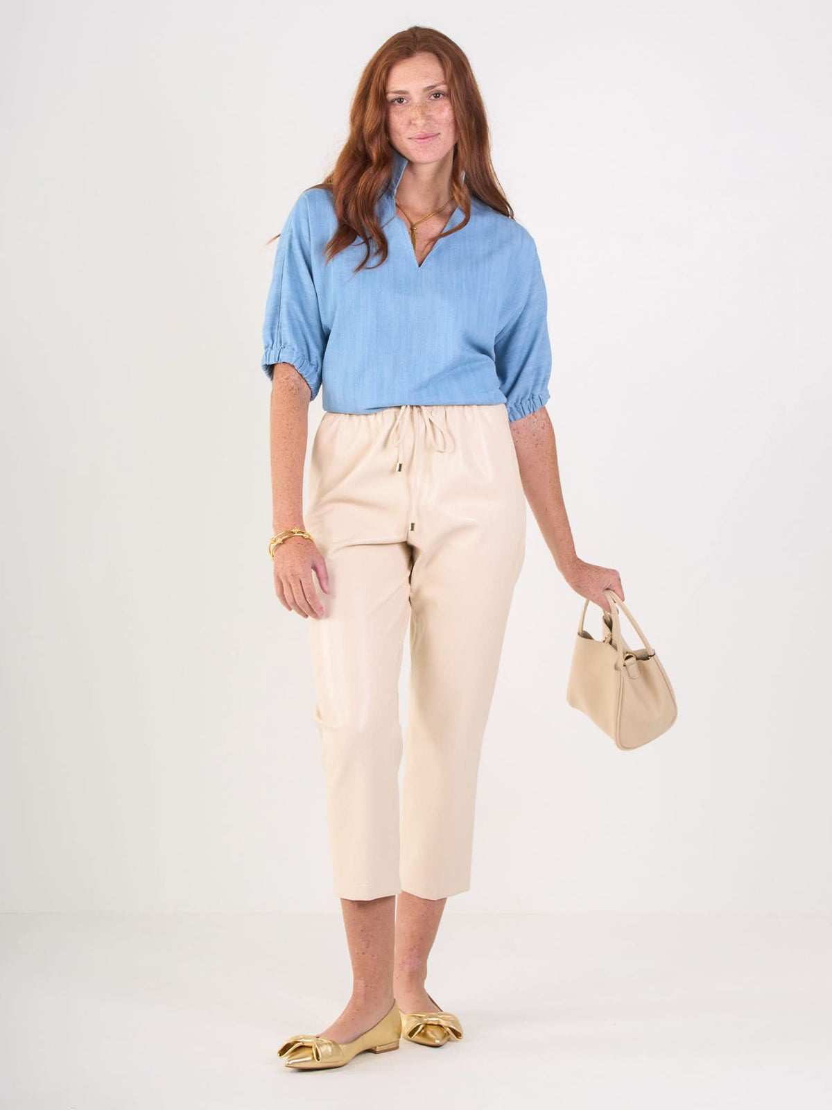 A woman with long red hair wears a Poppy Top in light blue, beige cropped pants, gold flats, and holds a small beige handbag while standing against a plain white background.