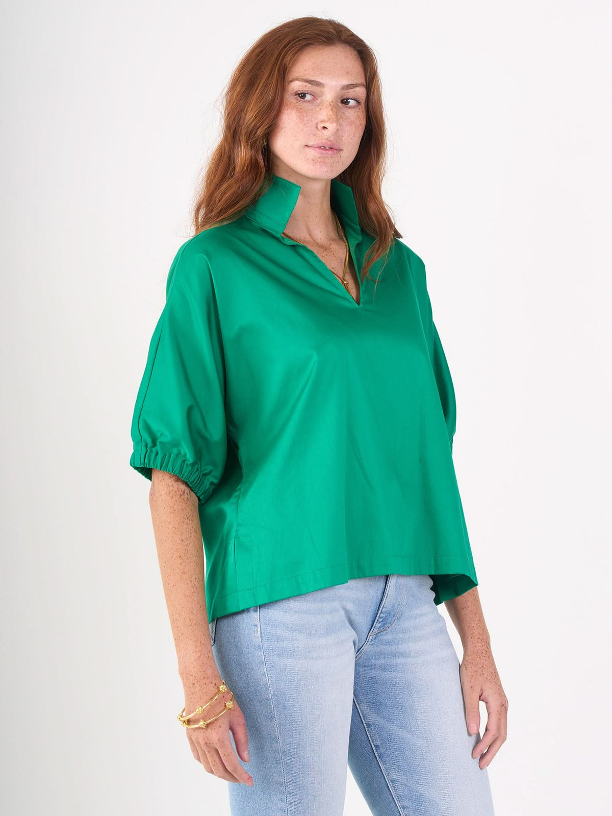 A woman with long red hair wears the Poppy Top in bright green cotton poplin and light blue jeans, standing against a plain white background with a neutral expression, looking slightly to the side.