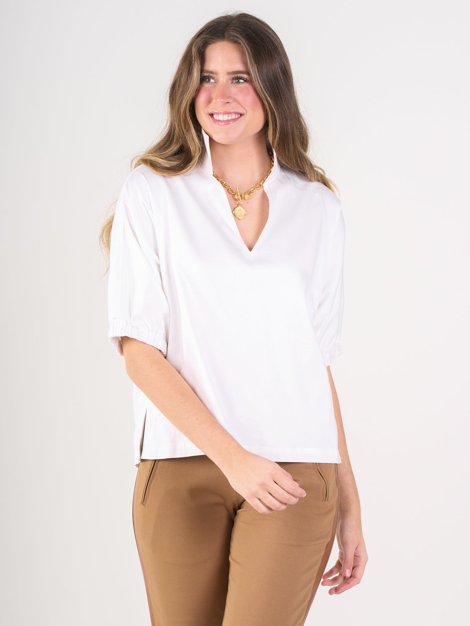 A woman with long brown hair is smiling against a plain light background, wearing the white cotton poplin Poppy Top featuring a V-neck and short puffed sleeves, paired with tan pants and a gold necklace.