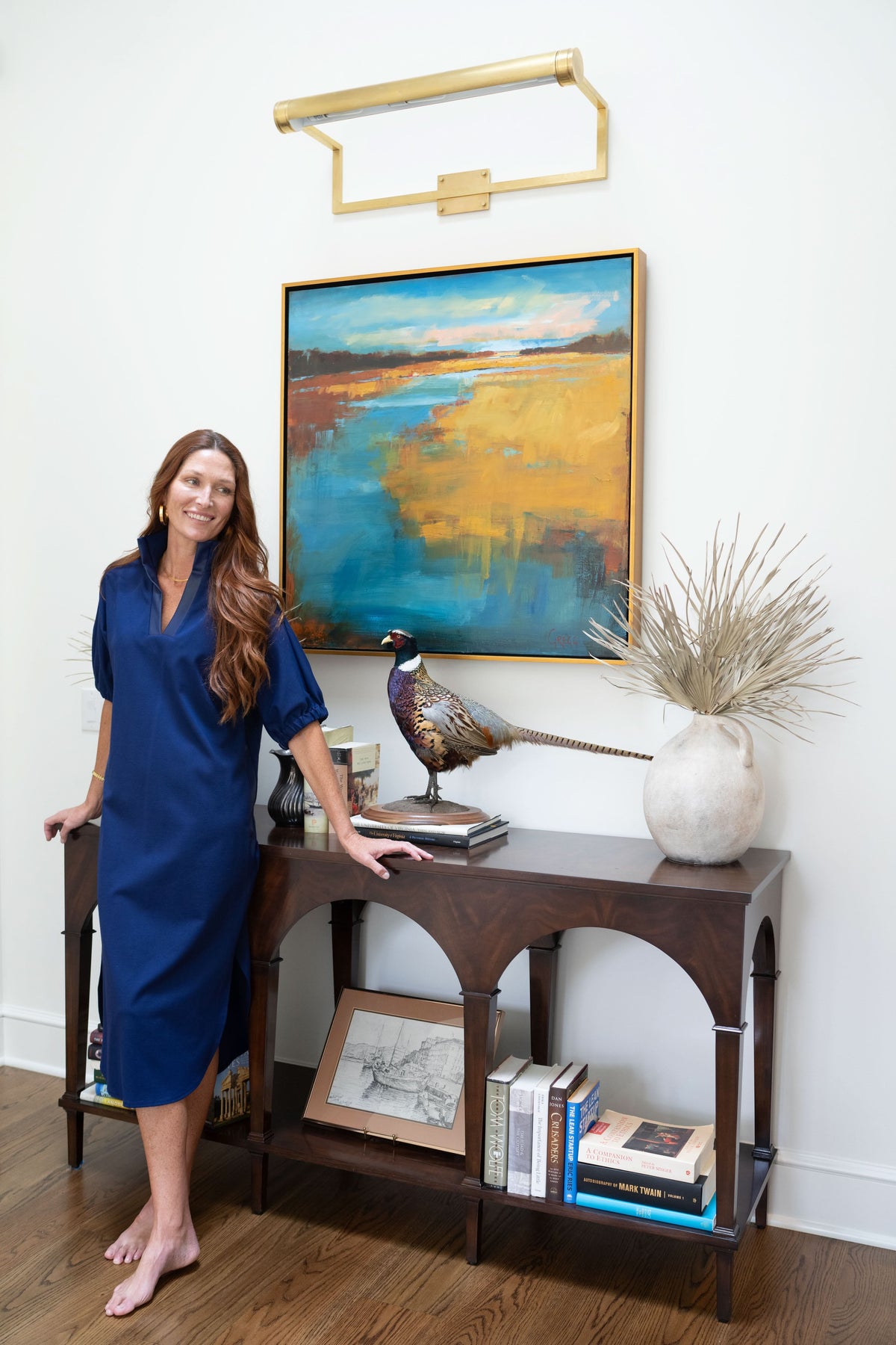 A woman in a navy ponte Poppy Caftan stands barefoot beside a dark wooden console table, which is adorned with books, a bird sculpture, a drawing, and a vase filled with dried foliage. Above the table hangs a vibrant landscape painting illuminated by a brass light fixture.