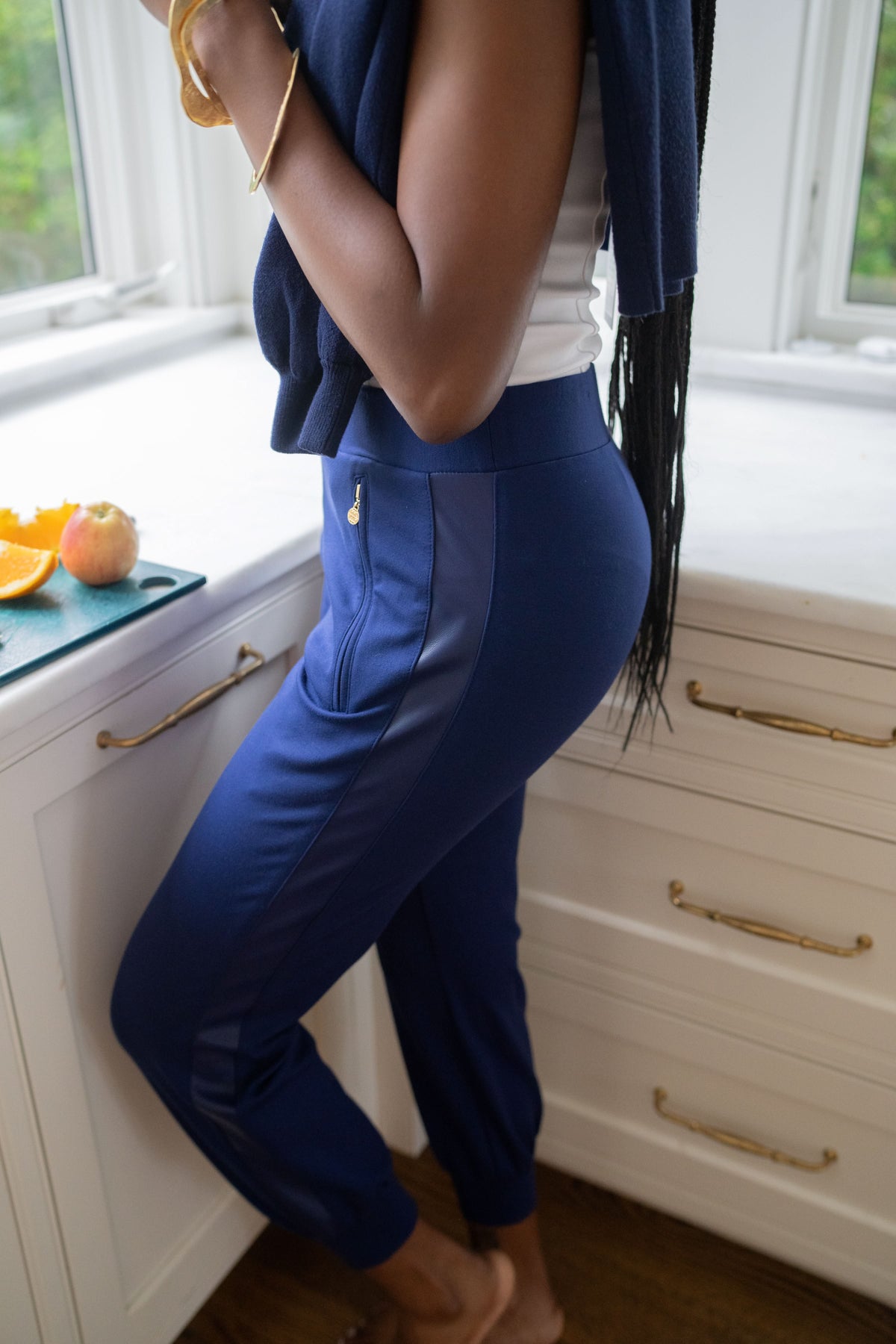A person dressed in a white tank top and the Downtown Jogger in navy ponte with supportive stretch stands in a kitchen by a counter containing sliced oranges and a peach. Their long, braided hair cascades over their shoulder as they hold a piece of fruit. The background showcases a window with lush greenery visible outside.