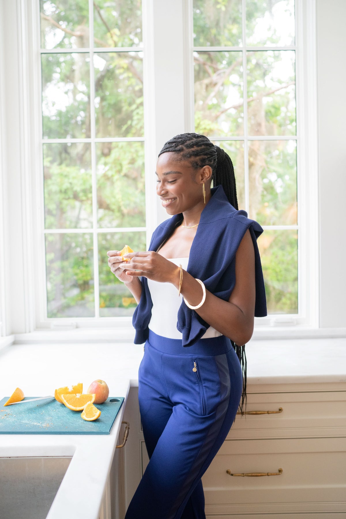 A woman with braided hair, dressed in the Navy Ponte Downtown Jogger and a blue top, stands by a kitchen counter with sliced oranges. She smiles while holding an orange slice, against the backdrop of large windows and trees.