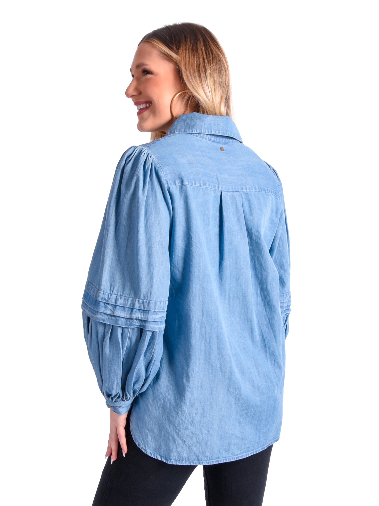 A woman with long hair is smiling, wearing the Oxford - Denim Chambray blouse featuring balloon sleeves and black pants, standing against a white background.