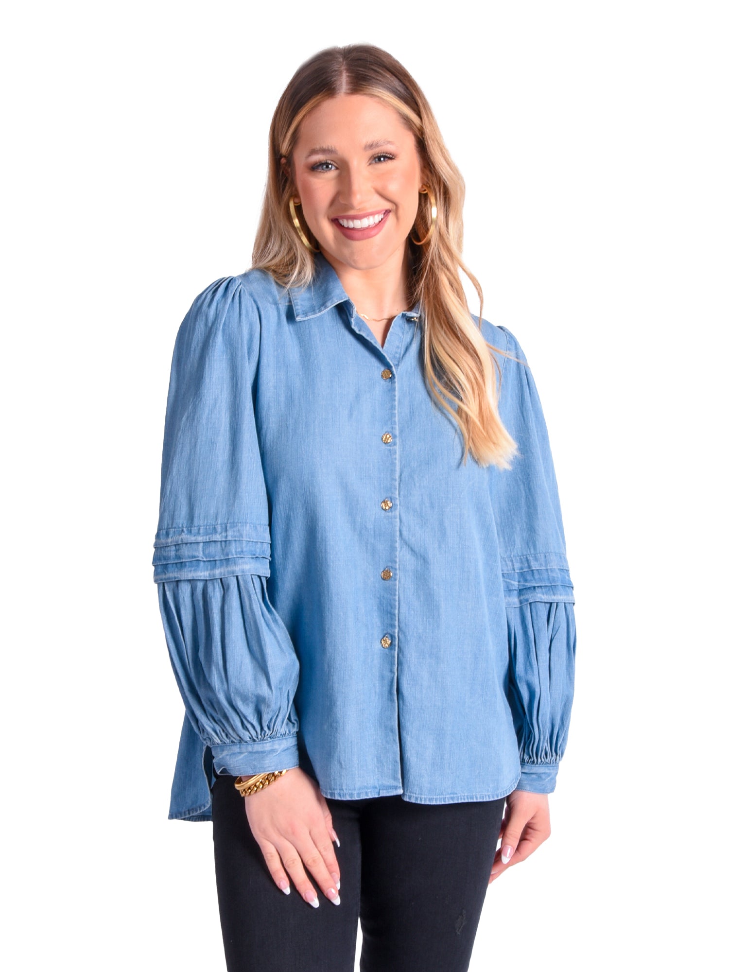 A smiling person with long hair stands against a white background, wearing the Oxford - Denim Chambray shirt featuring signature gold buttons and black pants. They accessorize with gold hoop earrings and a bracelet, completing their stylish look.