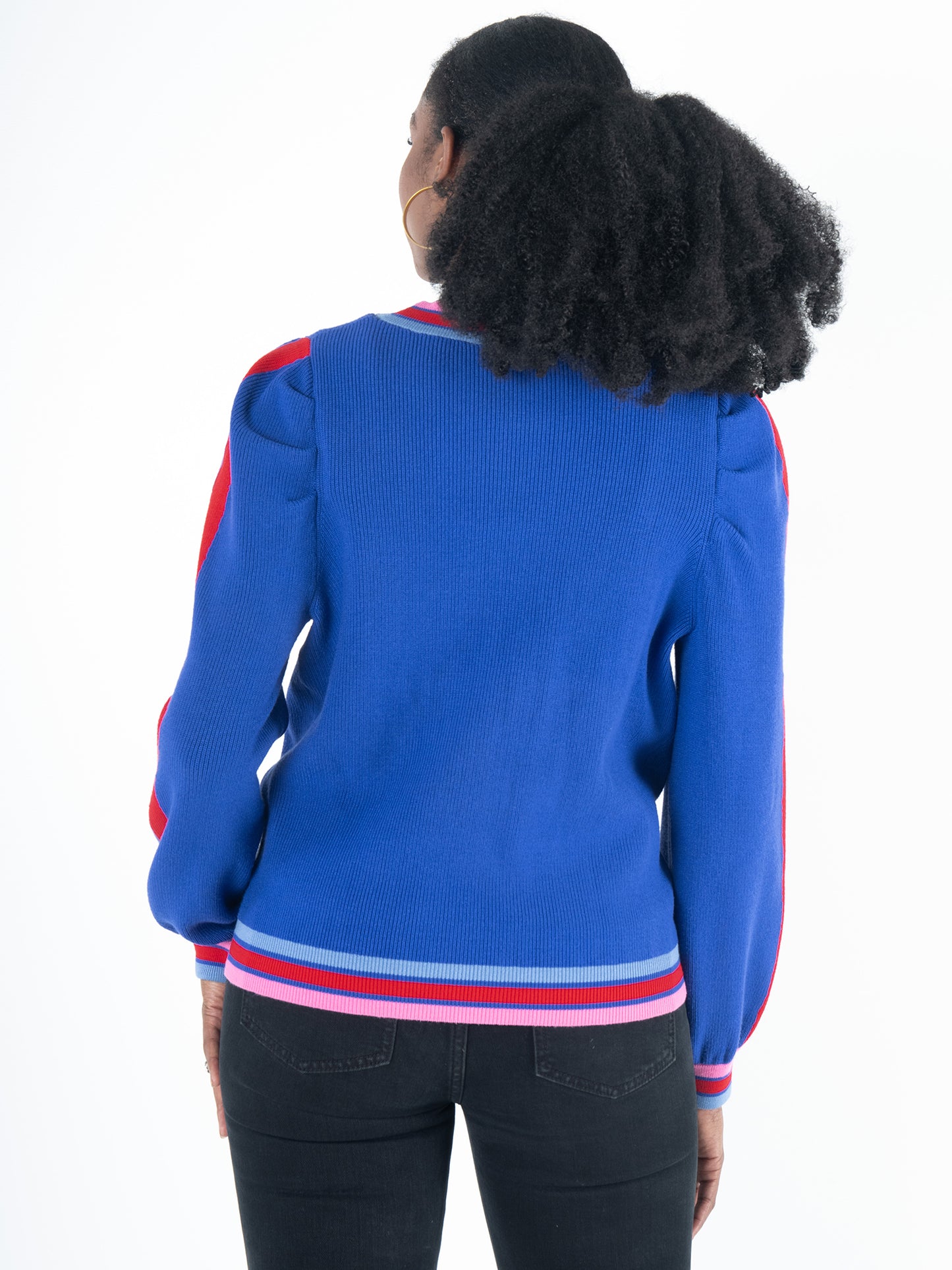 A woman with curly hair, wearing the chic Clara Cardigan - Bauhaus Stripe, faces away. The cardigan in blue features red shoulder accents, striped cuffs and hem, stylish puff sleeves, and gold button closures. She pairs it with black pants against a plain white background.