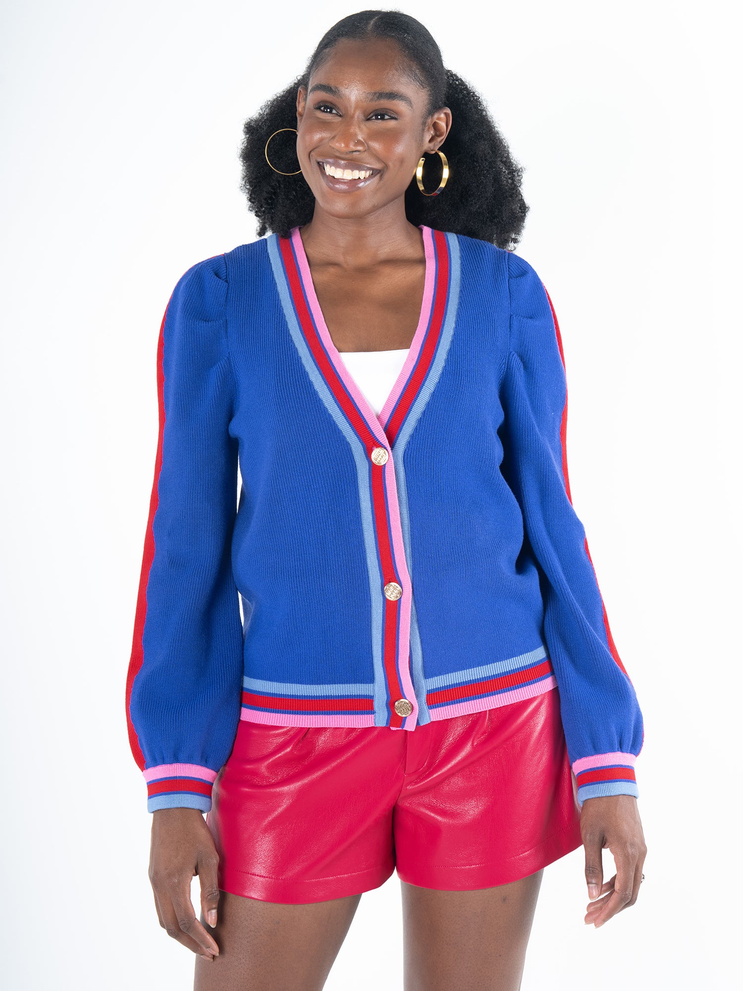 The woman stylishly dons the Clara Cardigan - Bauhaus Stripe, characterized by gold button closures and puff sleeves. Her outfit is composed of a blue cardigan with red stripes and pink trim layered over a white top, complemented beautifully by red shorts. She smiles warmly, her look further accentuated by hoop earrings against a plain background.