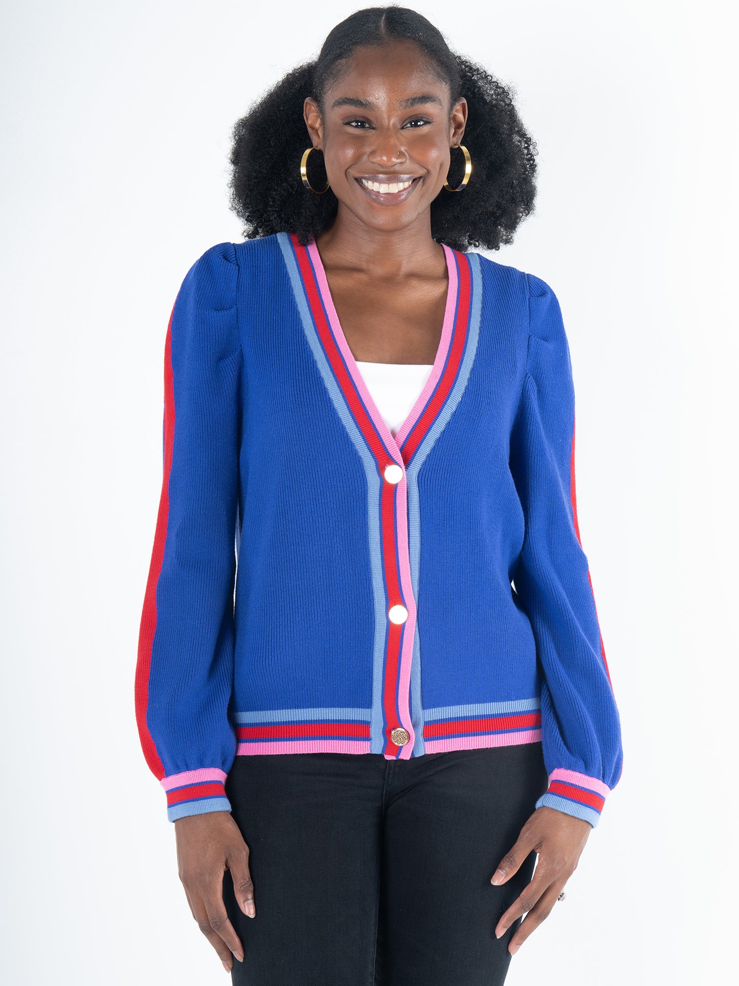 A smiling person wears the Clara Cardigan - Bauhaus Stripe, which features puff sleeves and gold button closures in its blue design with pink and red trim over a white top. Paired beautifully with black pants and gold hoop earrings, the backdrop is a simple white.