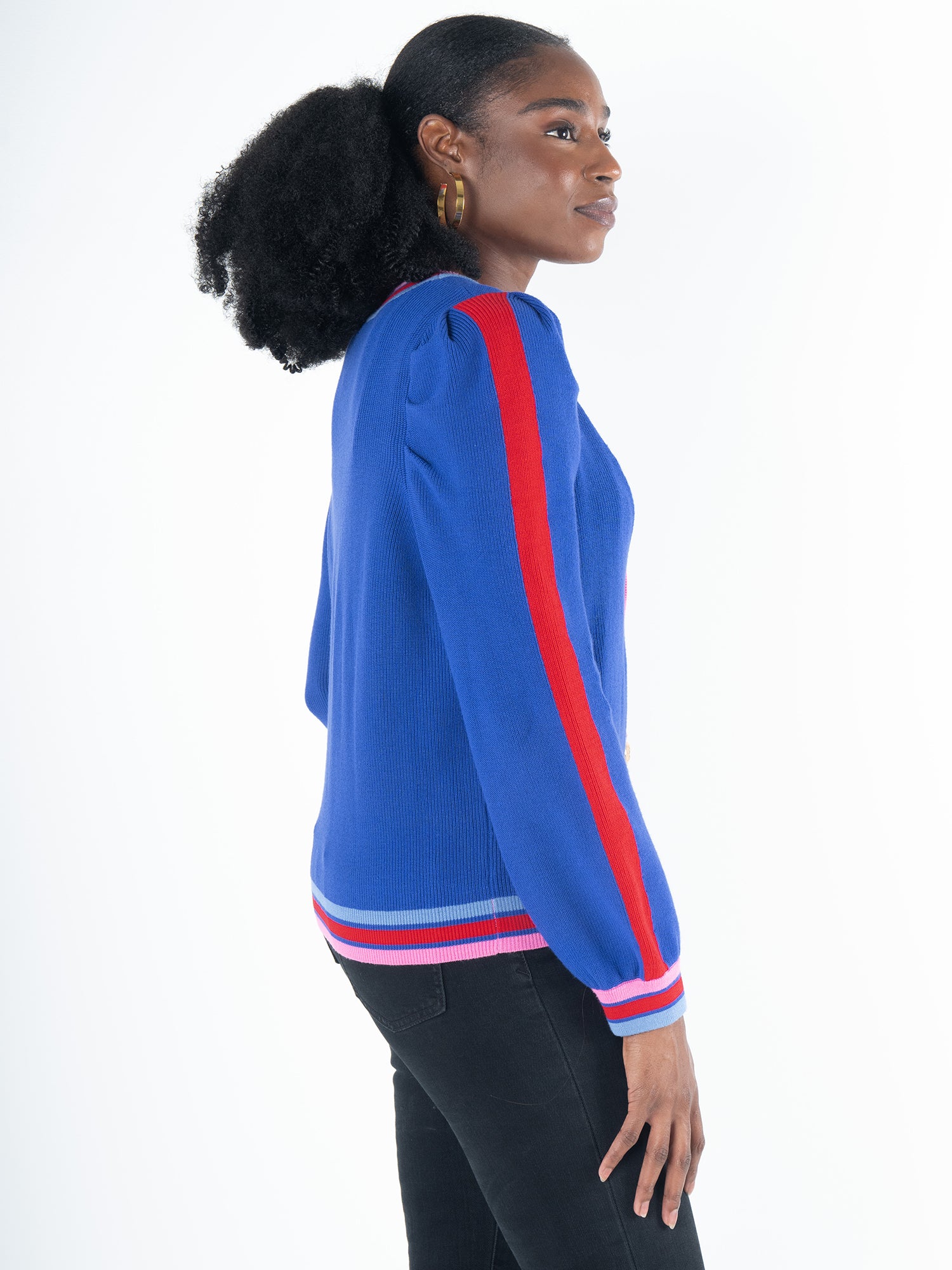 A person with curly hair is wearing the Clara Cardigan - Bauhaus Stripe in bright blue, showcasing red and pink striped sleeves and hem. The puff sleeves add a distinctive flair as they pose sideways against a plain white background. Gold button closures enhance the stylish appearance.