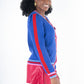 A person wearing the Clara Cardigan - Bauhaus Stripe with royal blue puff sleeves and gold button closures, paired with red shorts. They are smiling and standing in a side pose against a plain white background.