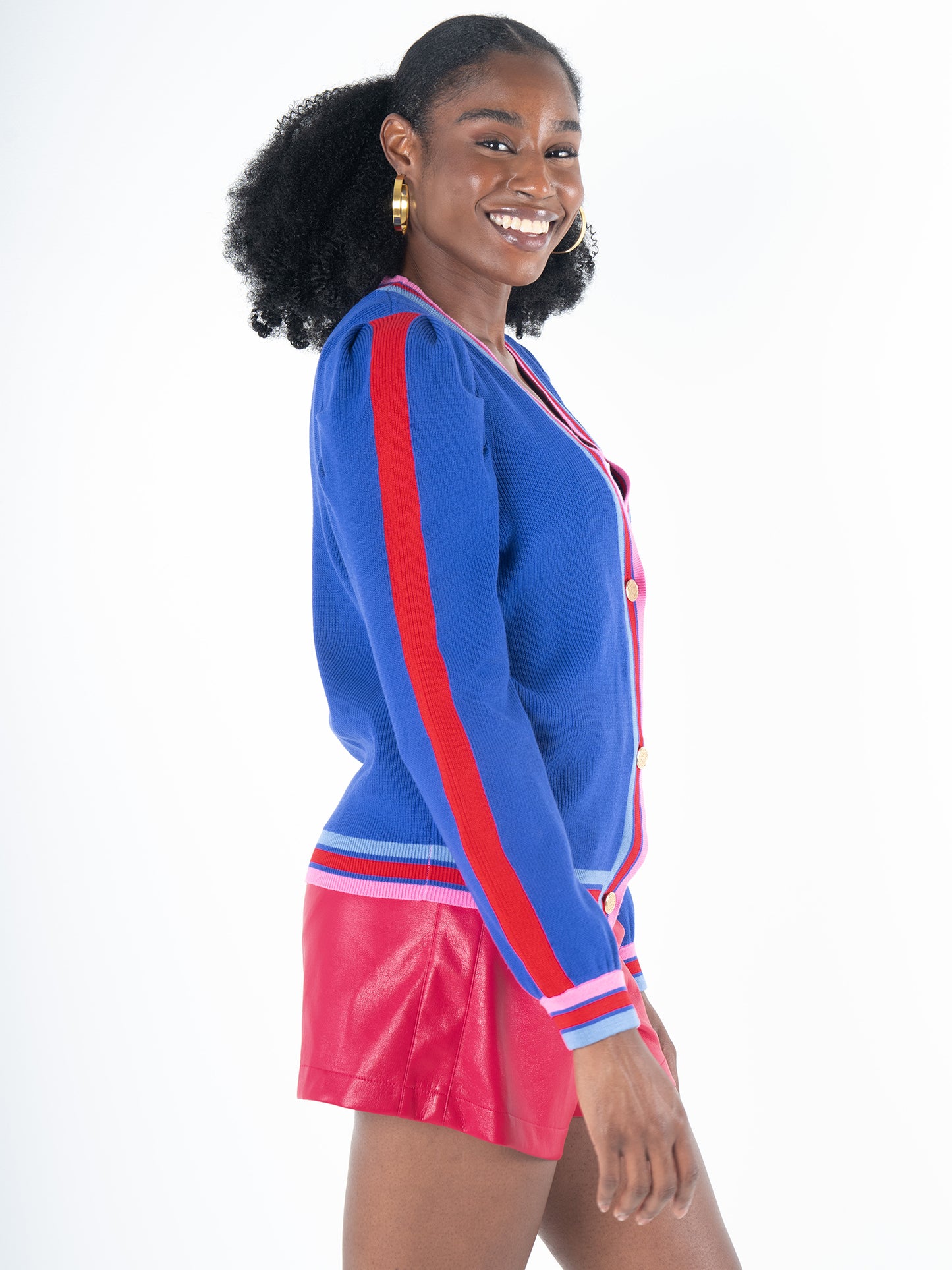 A person wearing the Clara Cardigan - Bauhaus Stripe with royal blue puff sleeves and gold button closures, paired with red shorts. They are smiling and standing in a side pose against a plain white background.