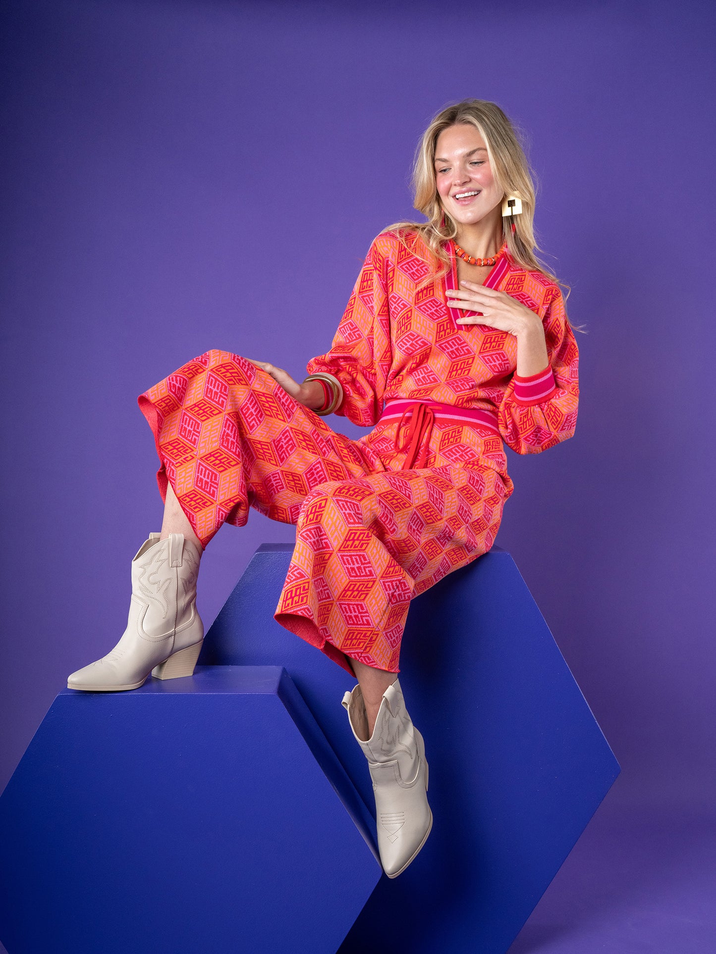 A person sits on a blue geometric shape, dressed in the Teddy Crop Trouser - Block Monogram, a vibrant red and pink patterned viscose knit ensemble with cropped wide-leg pants and white ankle boots. The solid purple backdrop highlights their smile as one hand gracefully rests near their chest.