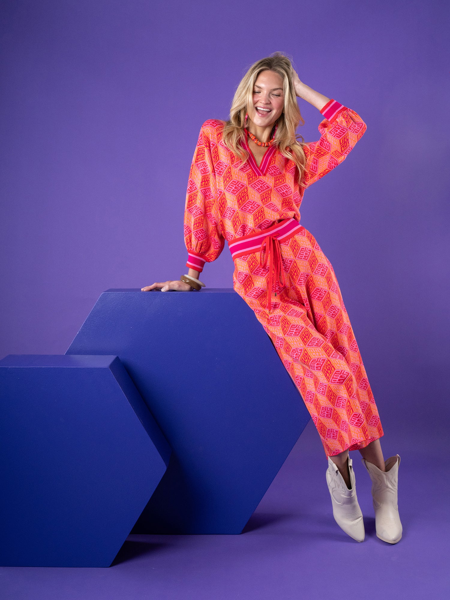 A woman in a vibrant pink and orange patterned outfit leans casually on a large blue geometric shape against a purple background. Her smile radiates confidence, complemented by white ankle boots and a pair of Teddy Crop Trousers - Block Monogram that add flair to her ensemble.