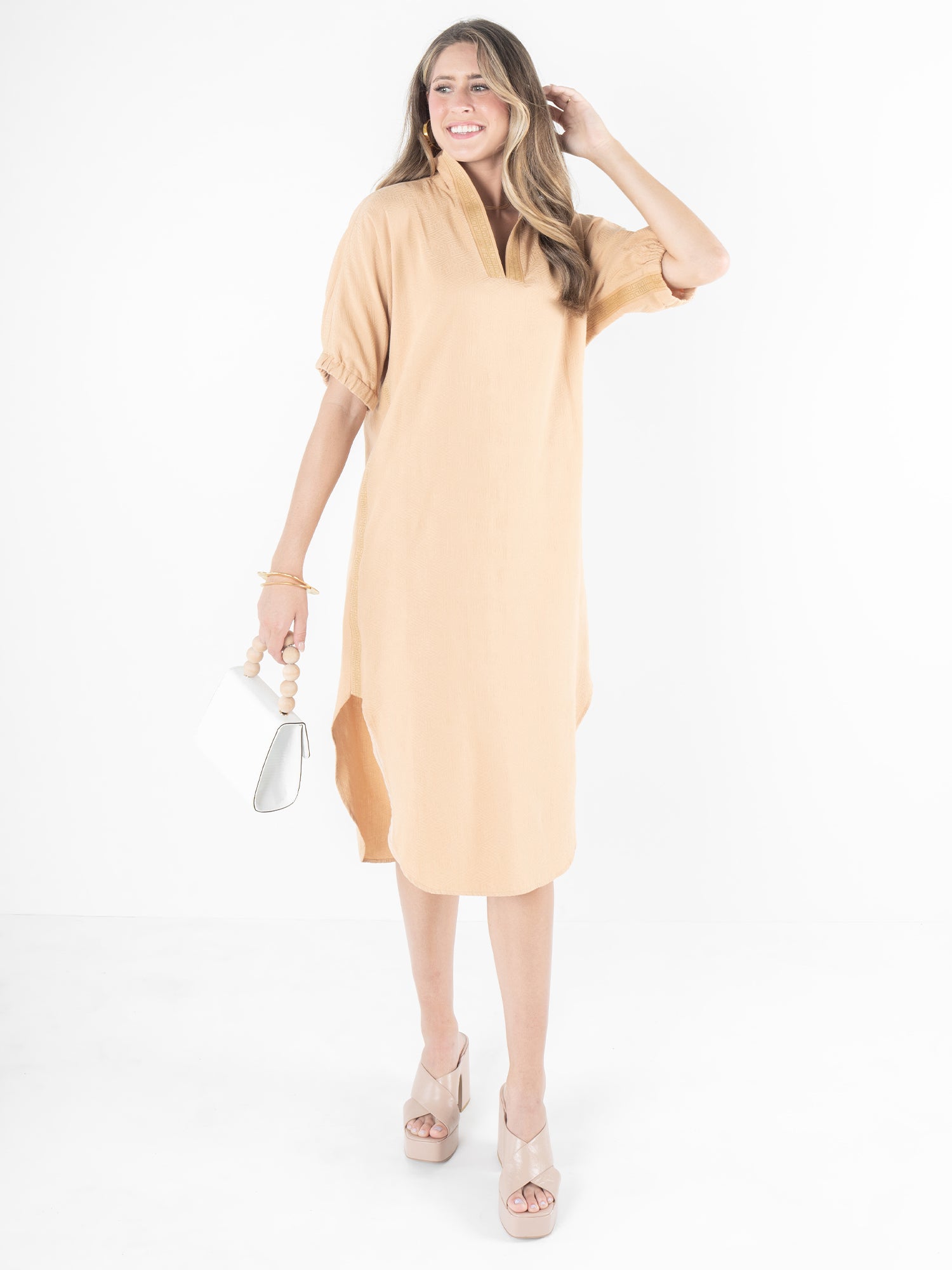 A woman in a peach dress stands smiling against a white background, holding a small white purse in her left hand and wearing beige platform sandals, highlighting the relaxed silhouette of the versatile piece known as the Poppy Caftan - Lark Monogram.