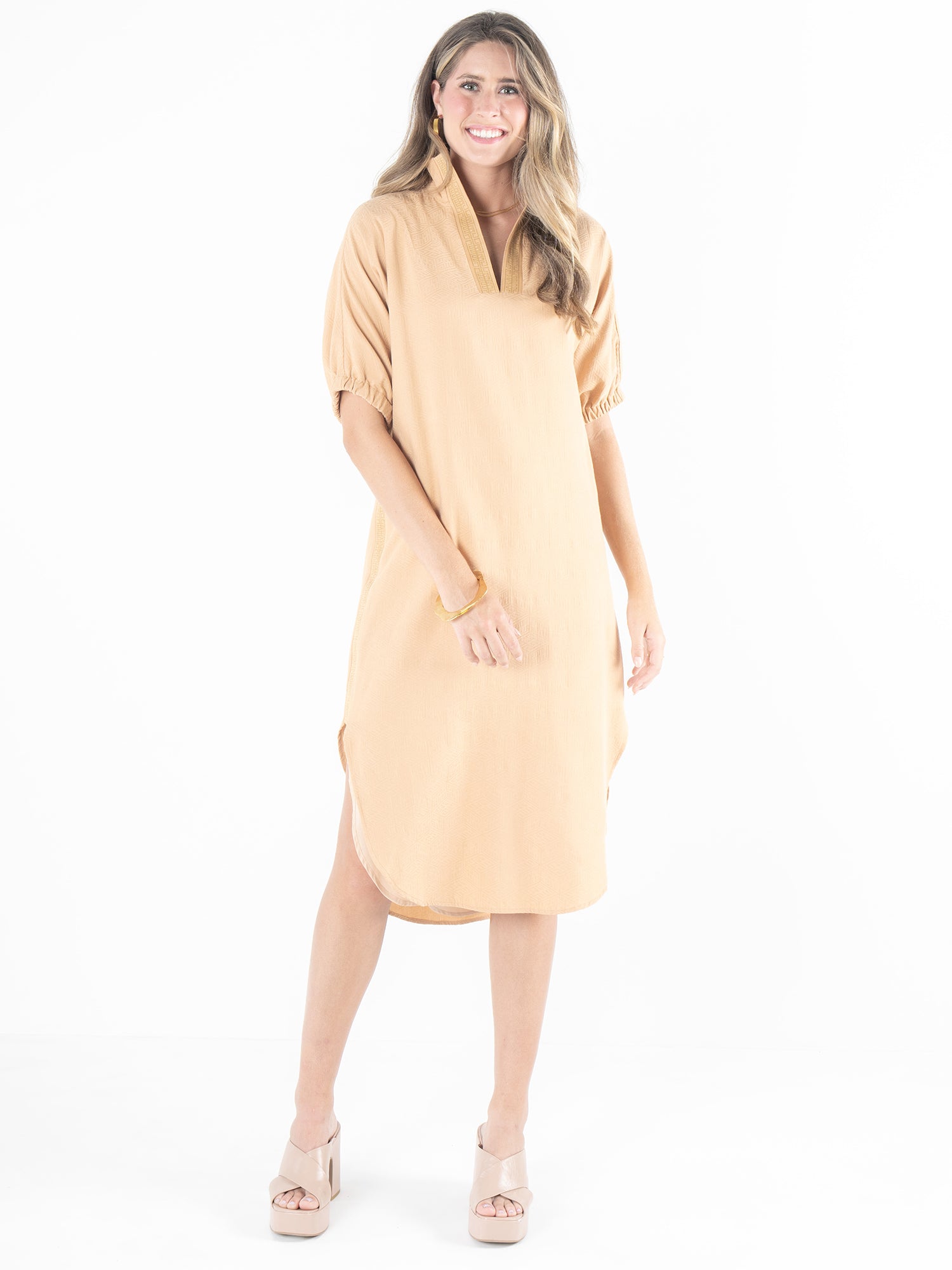 A person wearing the Poppy Caftan - Lark Monogram, a light tan, knee-length dress with puffed sleeves and open-toed heels, smiles against a white background, highlighting the garment's versatile and relaxed silhouette.