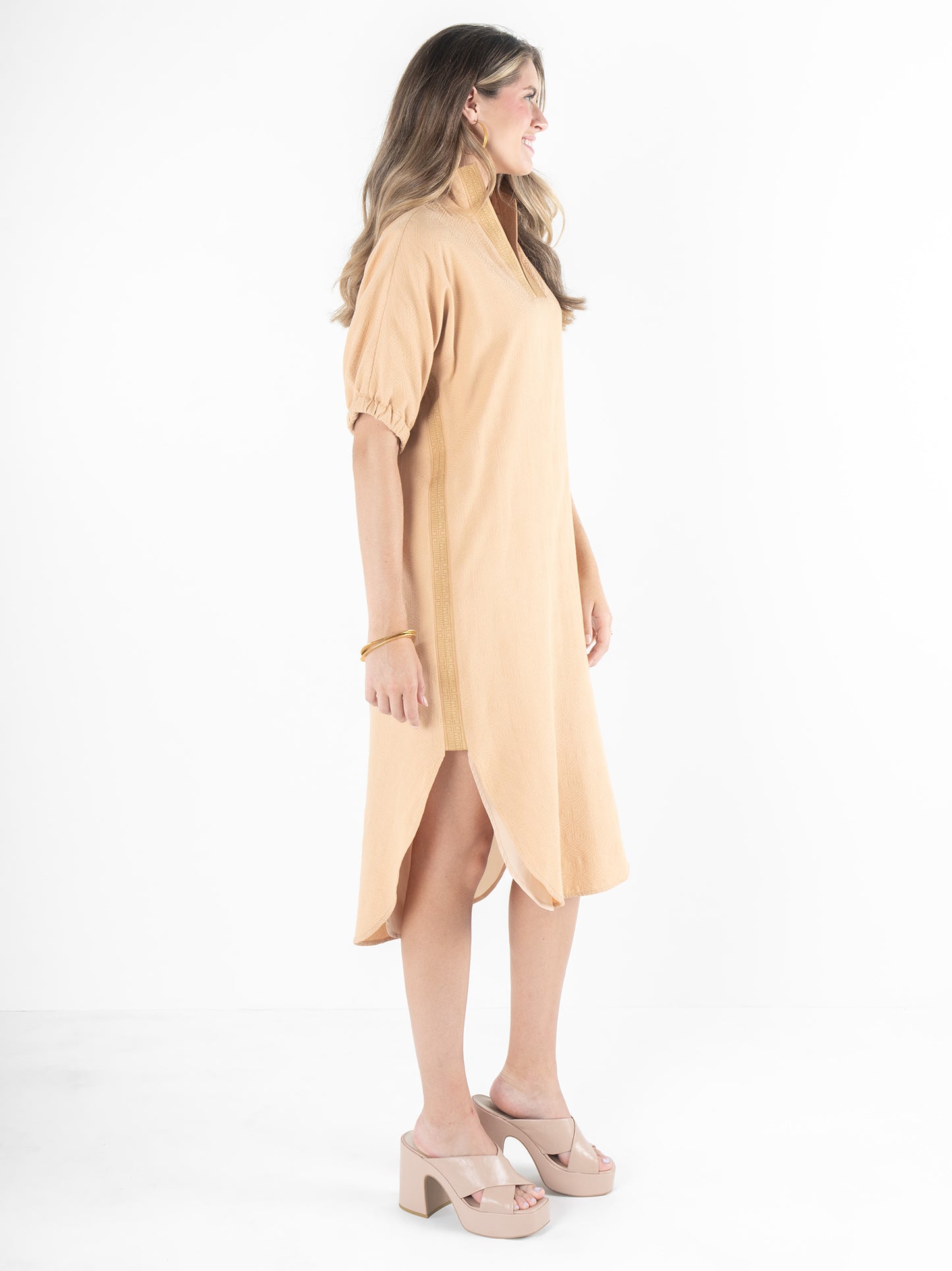 A woman in a beige, mid-length dress stands sideways against a plain white background. With long, wavy hair and platform sandals, she exudes effortless elegance. The relaxed silhouette of the Poppy Caftan - Lark Monogram features short sleeves and side slits for a truly versatile piece.