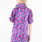 A person with long blond hair is seen standing away, showcasing the high collar and short sleeves of the vibrant Poppy Dress - Bauhaus, featuring a colorful print in shades of purple, pink, red, and blue.