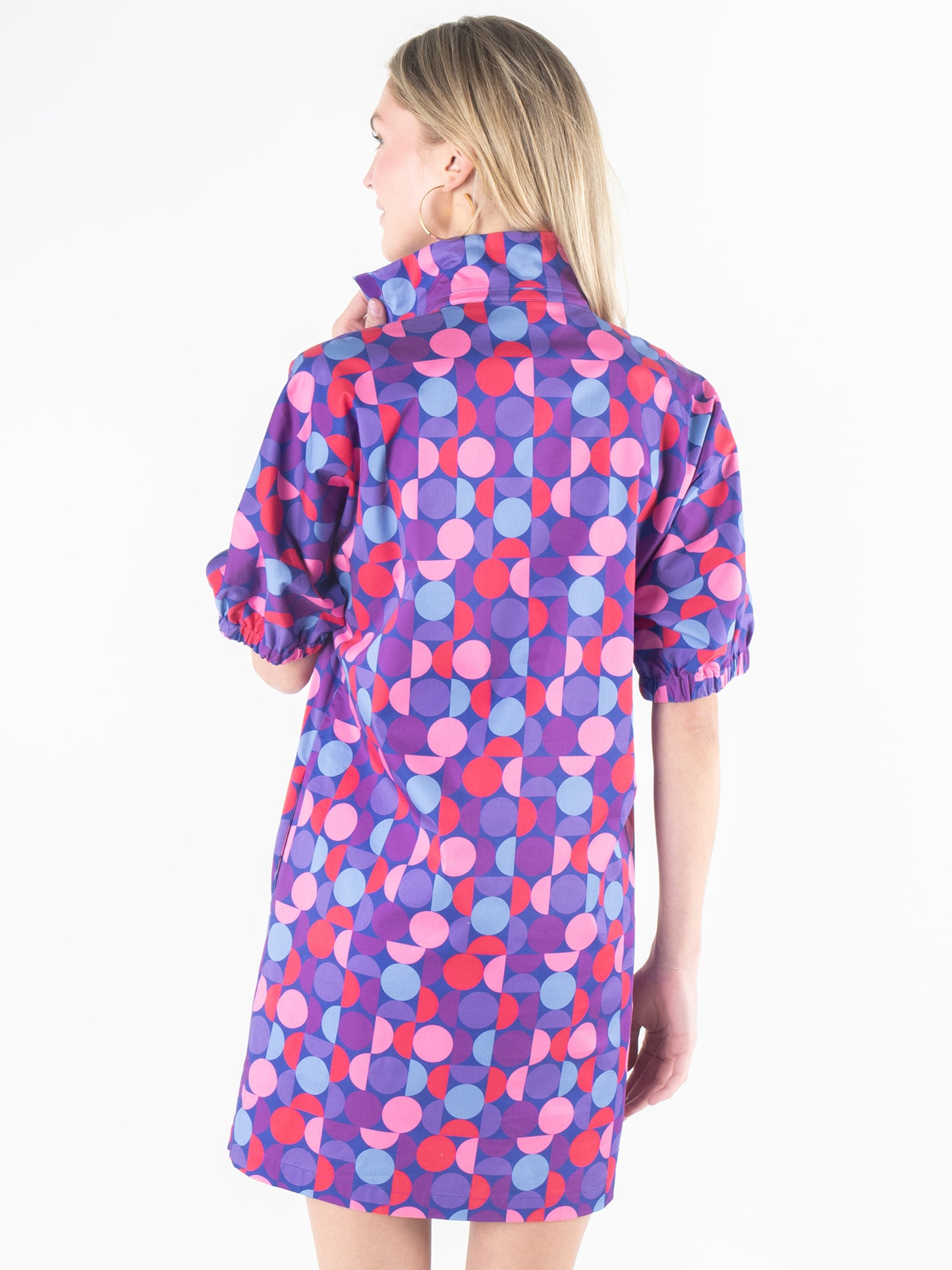 A person with long blond hair is seen standing away, showcasing the high collar and short sleeves of the vibrant Poppy Dress - Bauhaus, featuring a colorful print in shades of purple, pink, red, and blue.