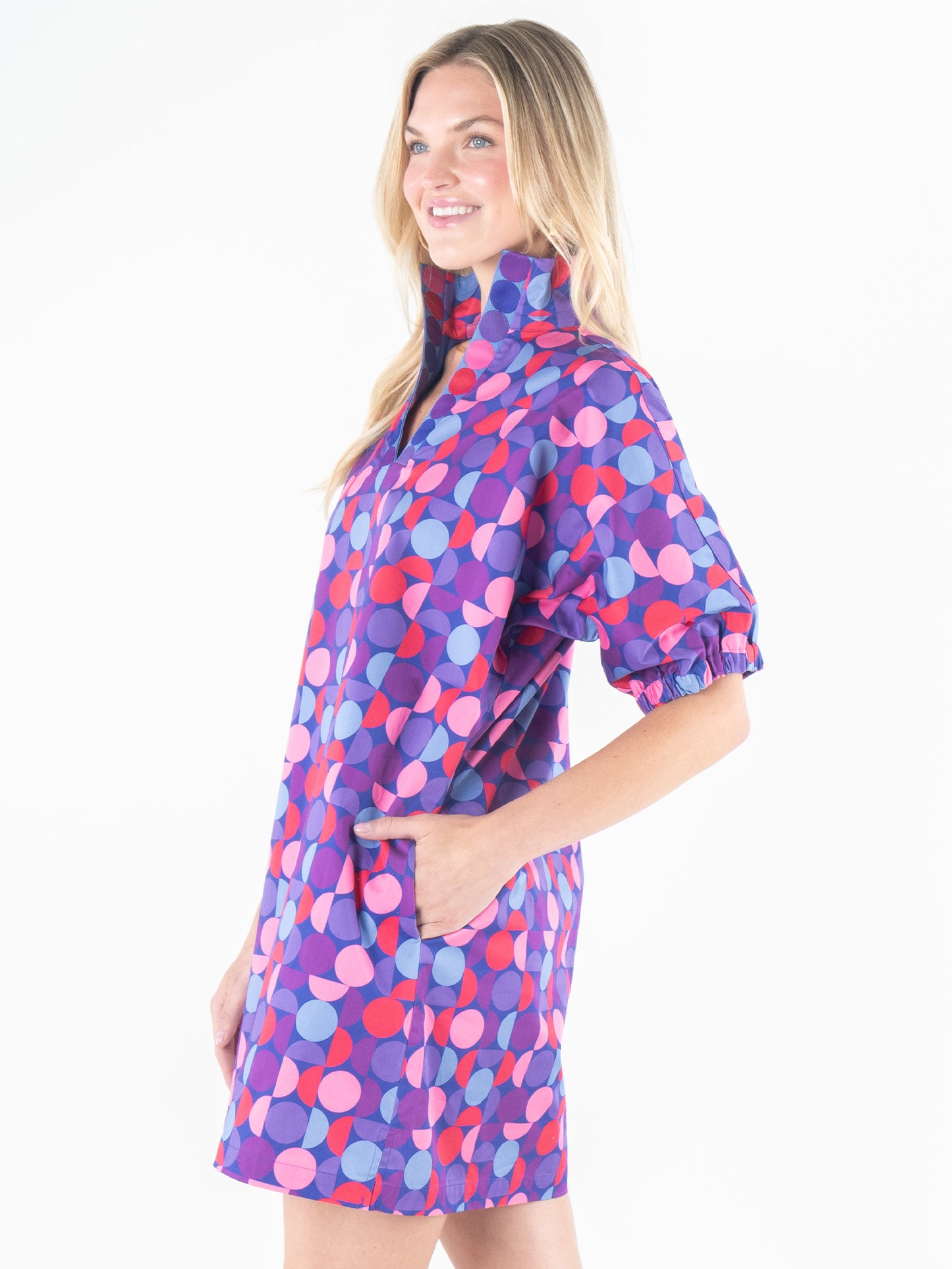 A woman wearing the Poppy Dress - Bauhaus, adorned with a vibrant mix of blue, purple, and pink circular patterns. This dress is distinguished by its short puff sleeves and high collar. With her hand in one pocket, she smiles as her long blonde hair flows over her shoulders.