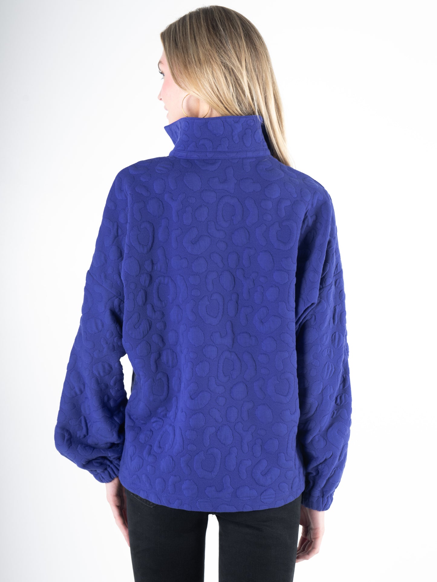 A woman with long blonde hair wears the Poppy Pop-Over in Royal Blue Cheetah, featuring a high collar and embossed fabric. She stands with her back to the camera against a plain white background, her long sleeves adding elegance to the ensemble.