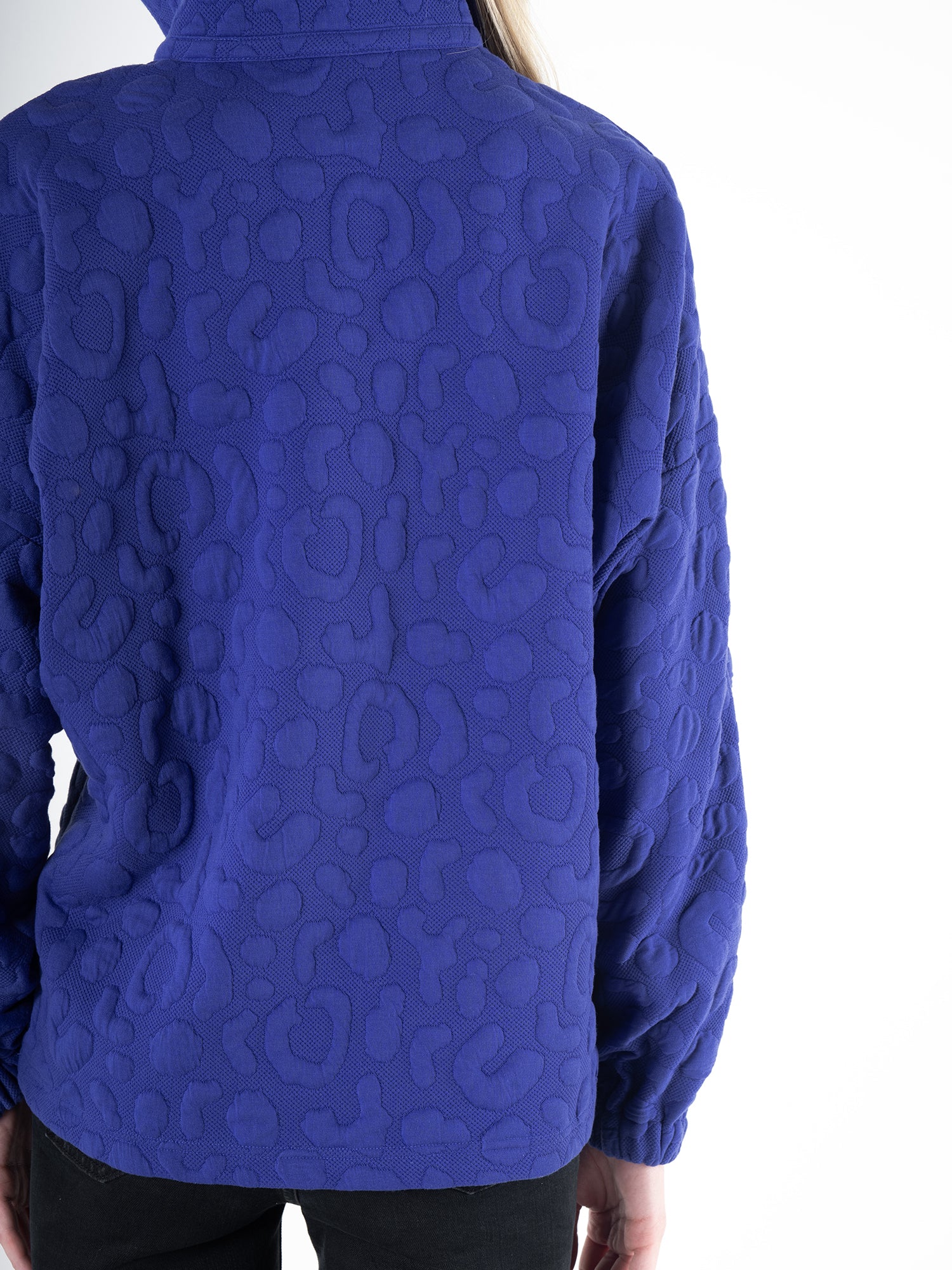 A person is facing away from the camera, wearing the Poppy Pop-Over in royal blue cheetah, a long sleeve textured patterned sweatshirt. Paired with black pants, they stand against a plain white backdrop.