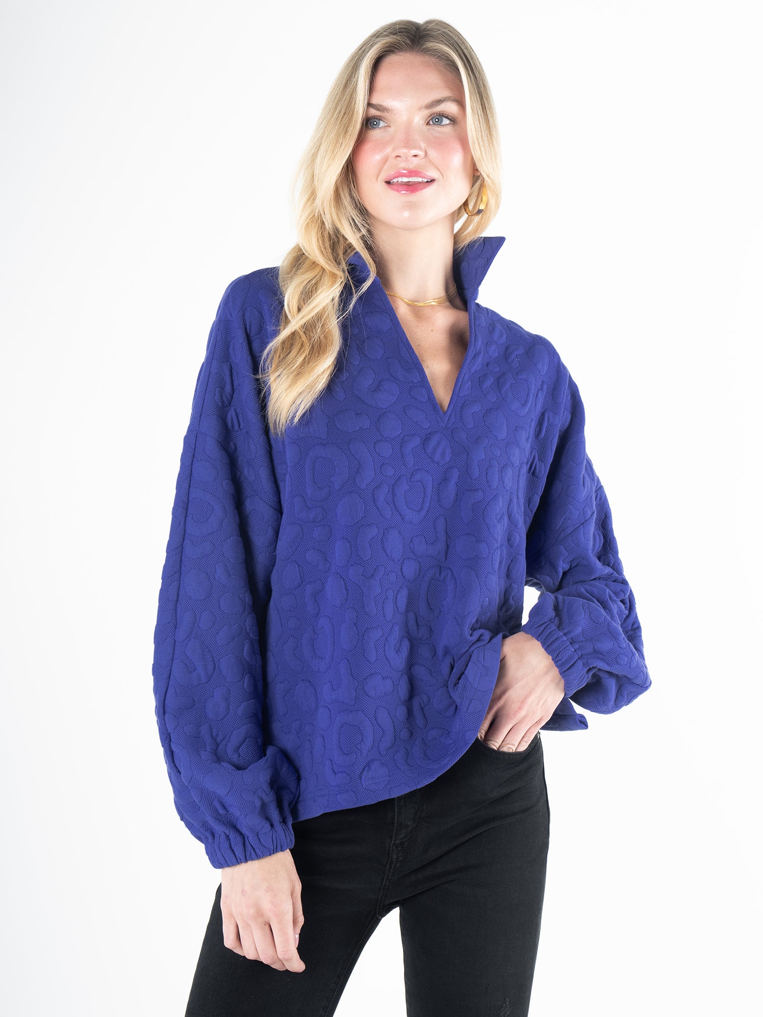 A woman in a Poppy Pop-Over Royal Blue Cheetah sweater with long sleeves and a high collar stands against a white background. Her black pants complement her long blonde hair, and she smiles slightly.