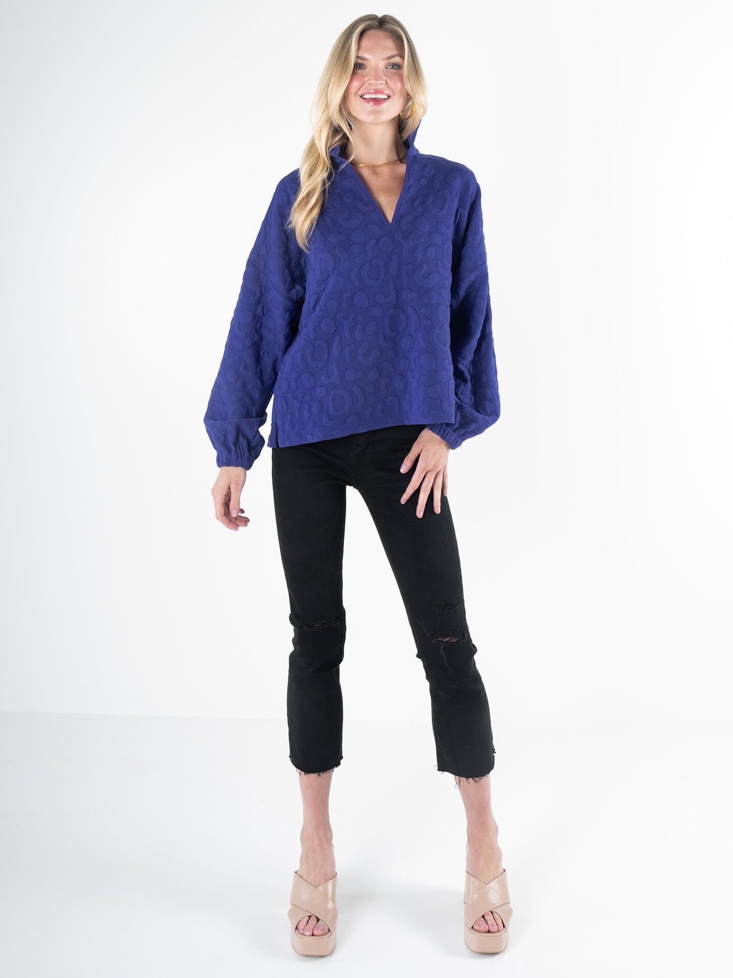 A woman with long blonde hair wears the Poppy Pop-Over in Royal Blue Cheetah, showcasing its puffy sleeves and embossed cheetah fabric. She pairs it with black distressed jeans and beige heeled sandals as she stands against a plain white background, smiling.