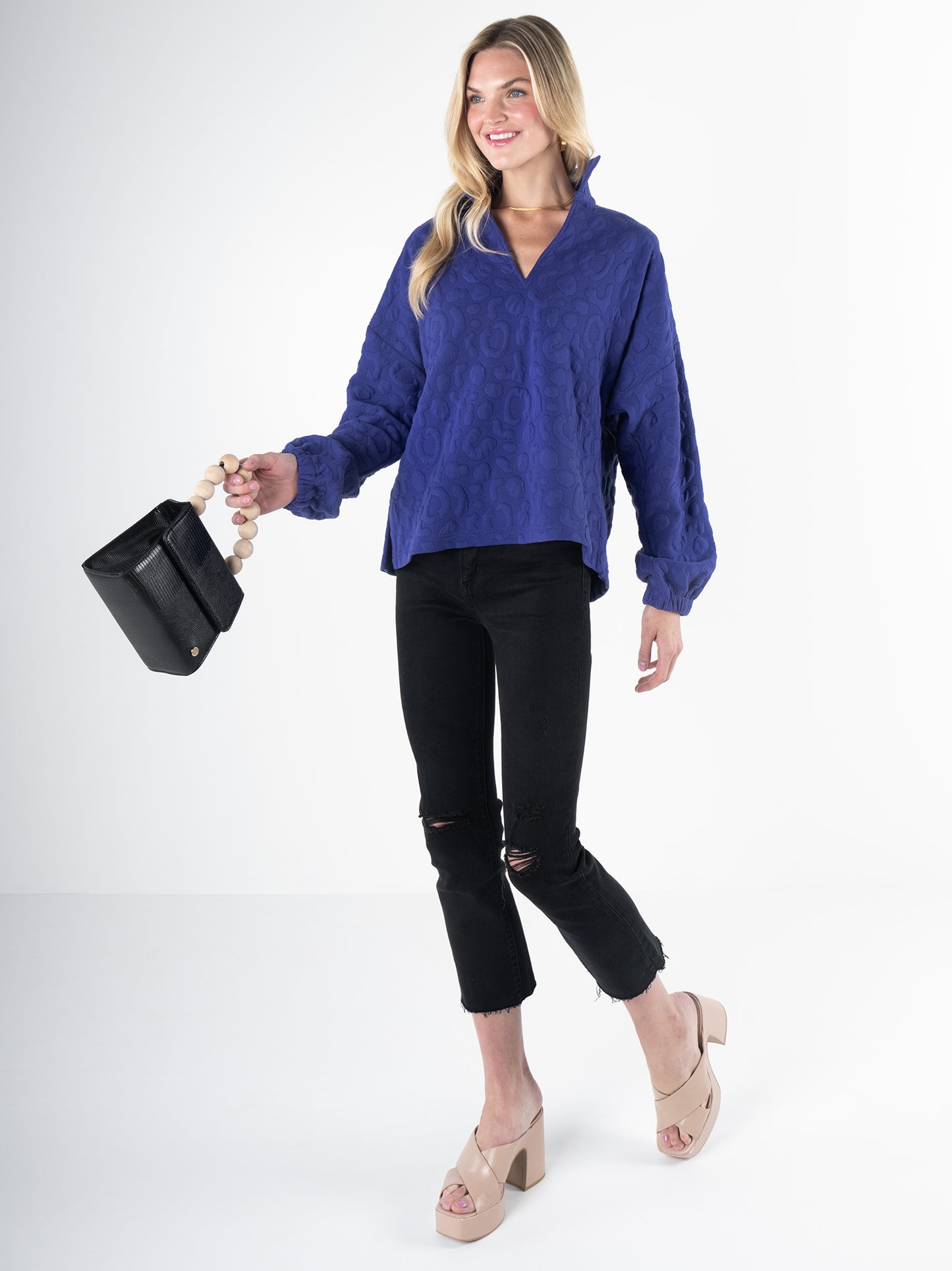 A person wearing a Poppy Pop-Over blouse in Royal Blue Cheetah and black ripped jeans holds a black handbag. They are wearing platform sandals and have long, blond hair against a plain white background.