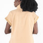 A person with curly hair styled in a ponytail is seen from the rear, dressed in the Poppy Pullover featuring the Lark Monogram. The beige short-sleeve top boasts a subtle pattern and folded sleeves and is paired with tan pants. They also accessorize with hoop earrings.