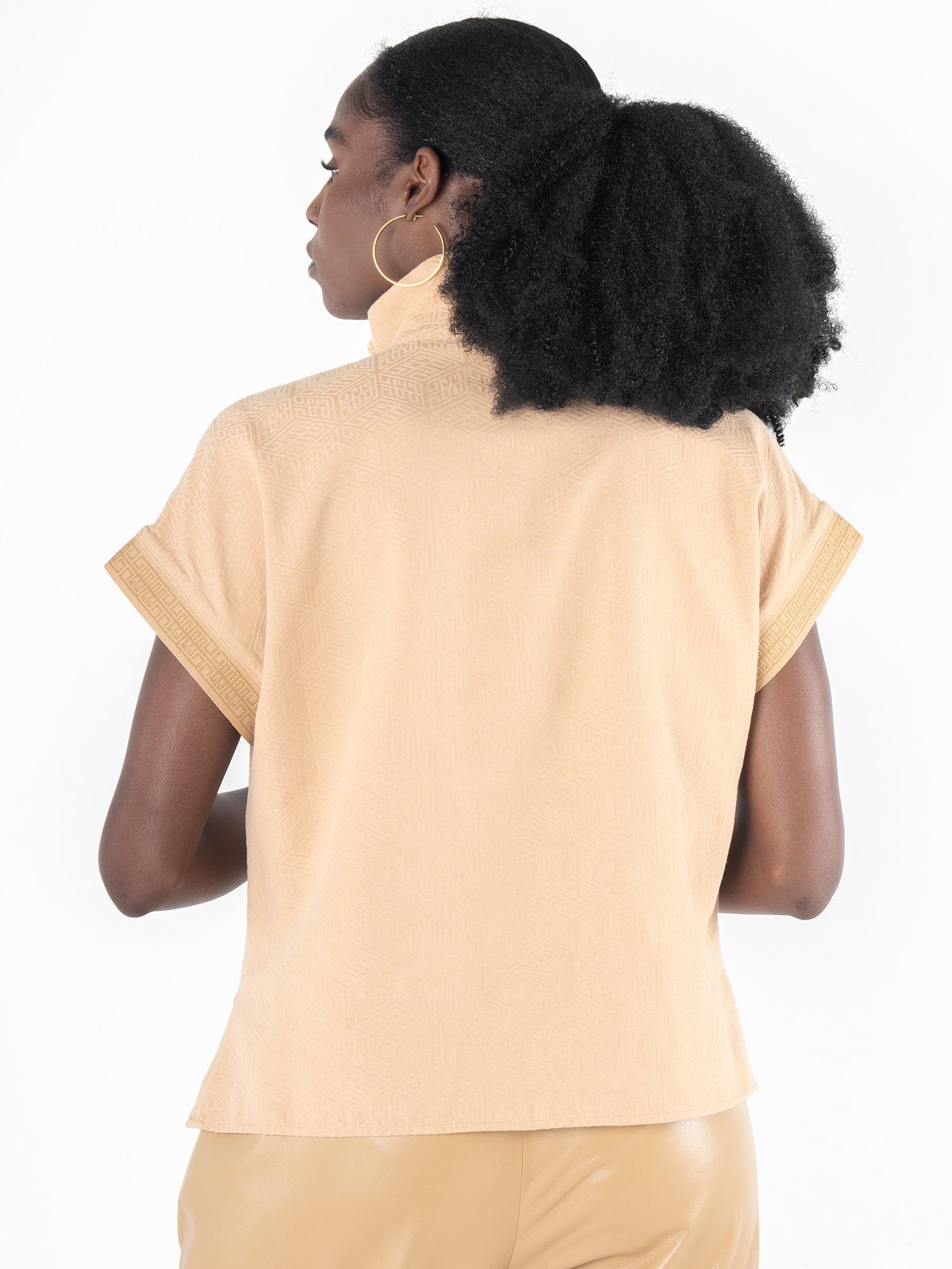 A person with curly hair styled in a ponytail is seen from the rear, dressed in the Poppy Pullover featuring the Lark Monogram. The beige short-sleeve top boasts a subtle pattern and folded sleeves and is paired with tan pants. They also accessorize with hoop earrings.