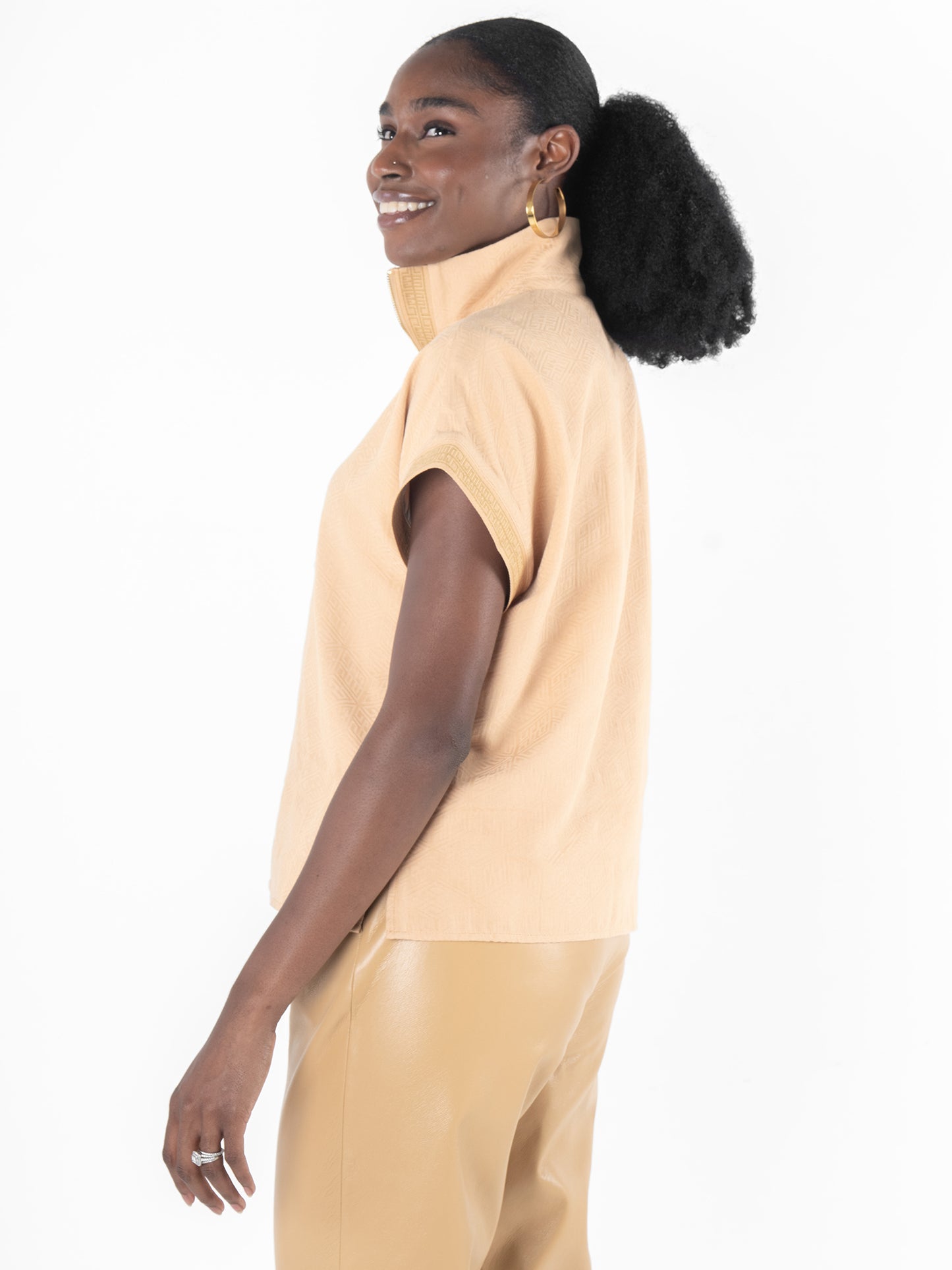 A woman with a ponytail smiles over her shoulder, wearing the Poppy Pullover - Lark Monogram sleeveless beige top with a high collar and matching pants. Her ensemble subtly showcases the Lark Monogram, adding an elegant touch against the plain white background.