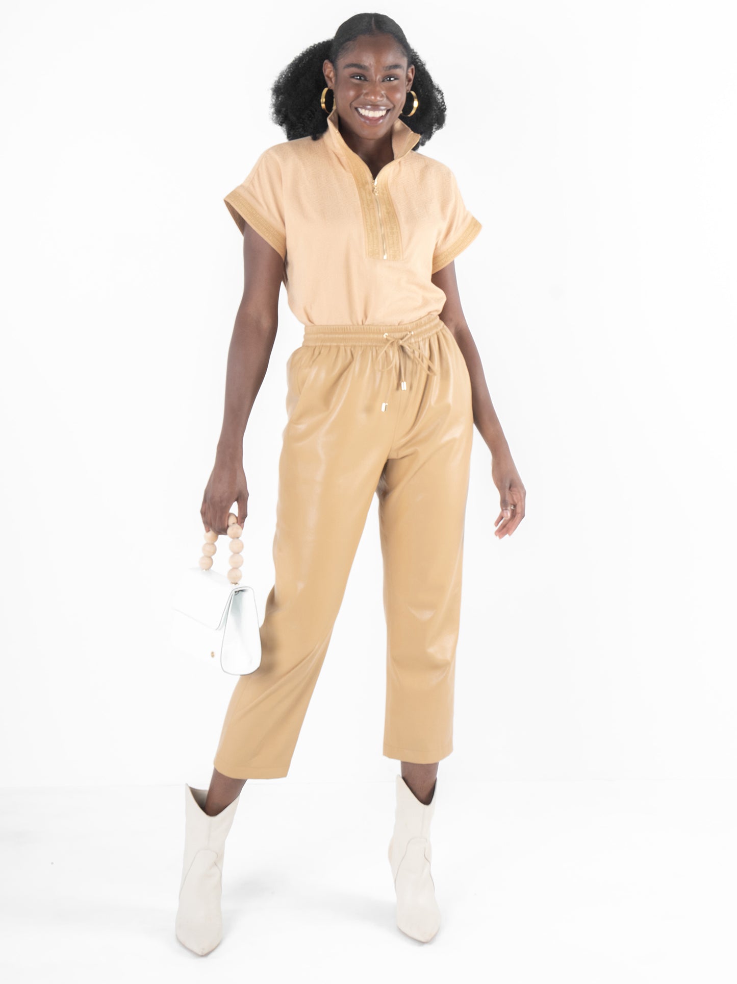 A person wearing a Lark Monogram Poppy Pullover stands smiling in a light brown outfit with short sleeves and cropped pants. They hold a small white handbag featuring the Signature Monogram Logo and wear white ankle boots. Their hair is styled in voluminous curls.