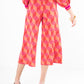 A person wearing the Block Monogram Teddy Crop Trouser featuring a geometric pattern in red and pink, paired with a matching top, is seen from the back. This viscose knit ensemble includes long sleeves and high block-heeled shoes, adding a modern twist to its cropped wide-leg design.