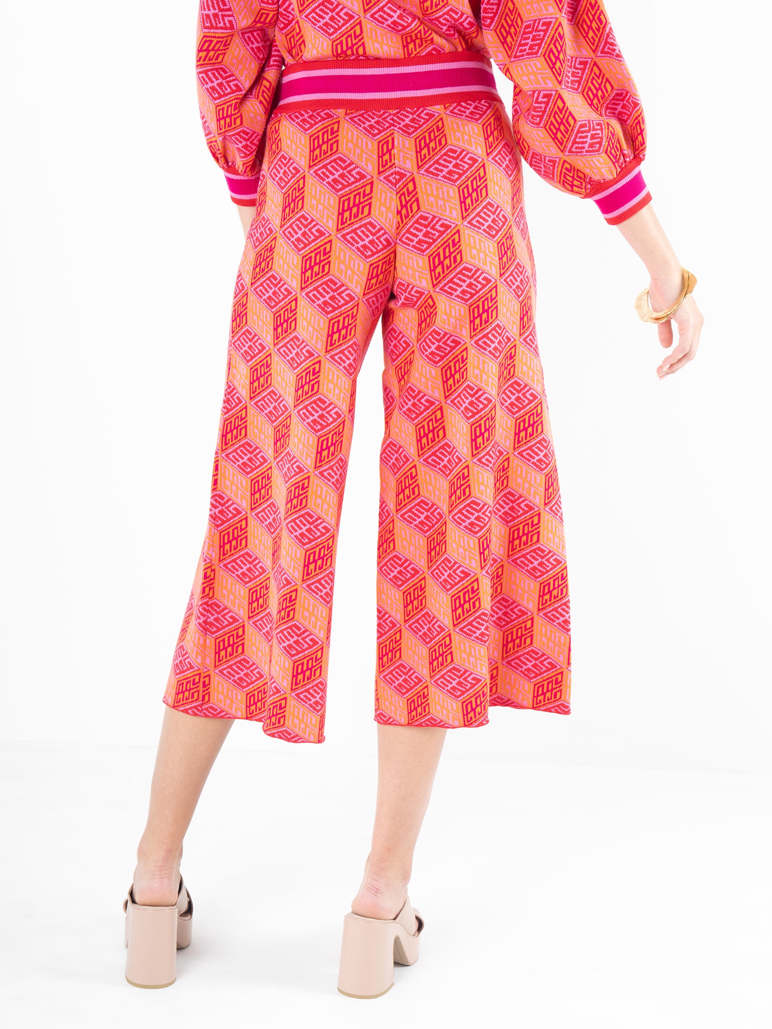 A person wearing the Block Monogram Teddy Crop Trouser featuring a geometric pattern in red and pink, paired with a matching top, is seen from the back. This viscose knit ensemble includes long sleeves and high block-heeled shoes, adding a modern twist to its cropped wide-leg design.