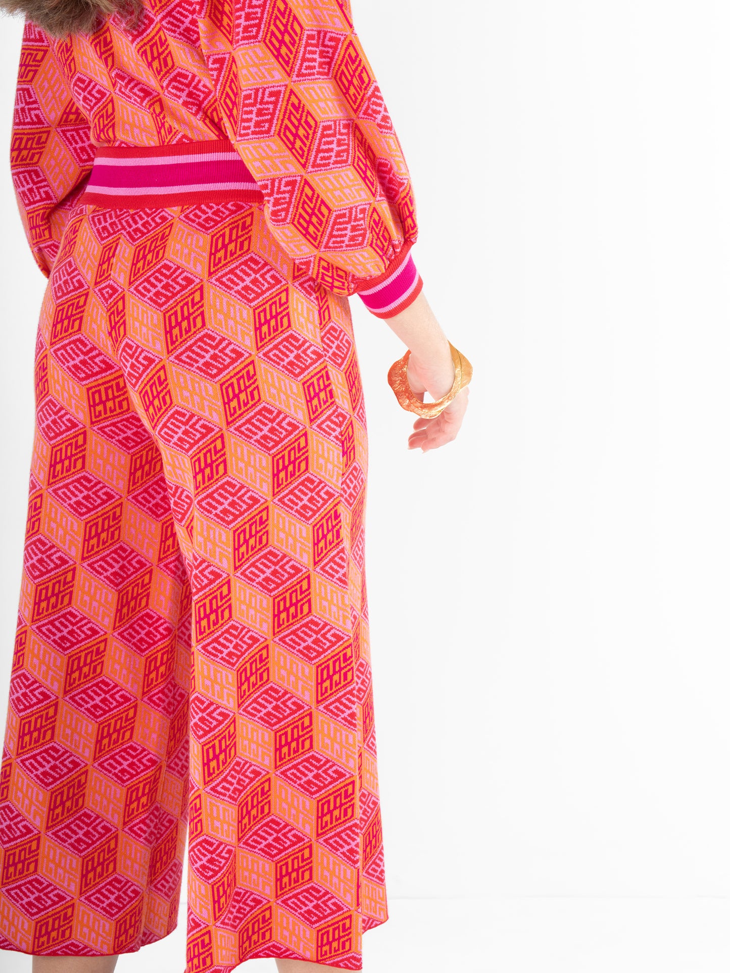 A person wearing the Teddy Crop Trouser - Block Monogram is dressed in a vibrant orange and pink geometric patterned outfit, with a long-sleeve top and cropped wide-leg pants. The viscose knit ensemble is highlighted by a pink and white striped waistband and cuffs, and they accessorize with a bracelet on their left wrist.