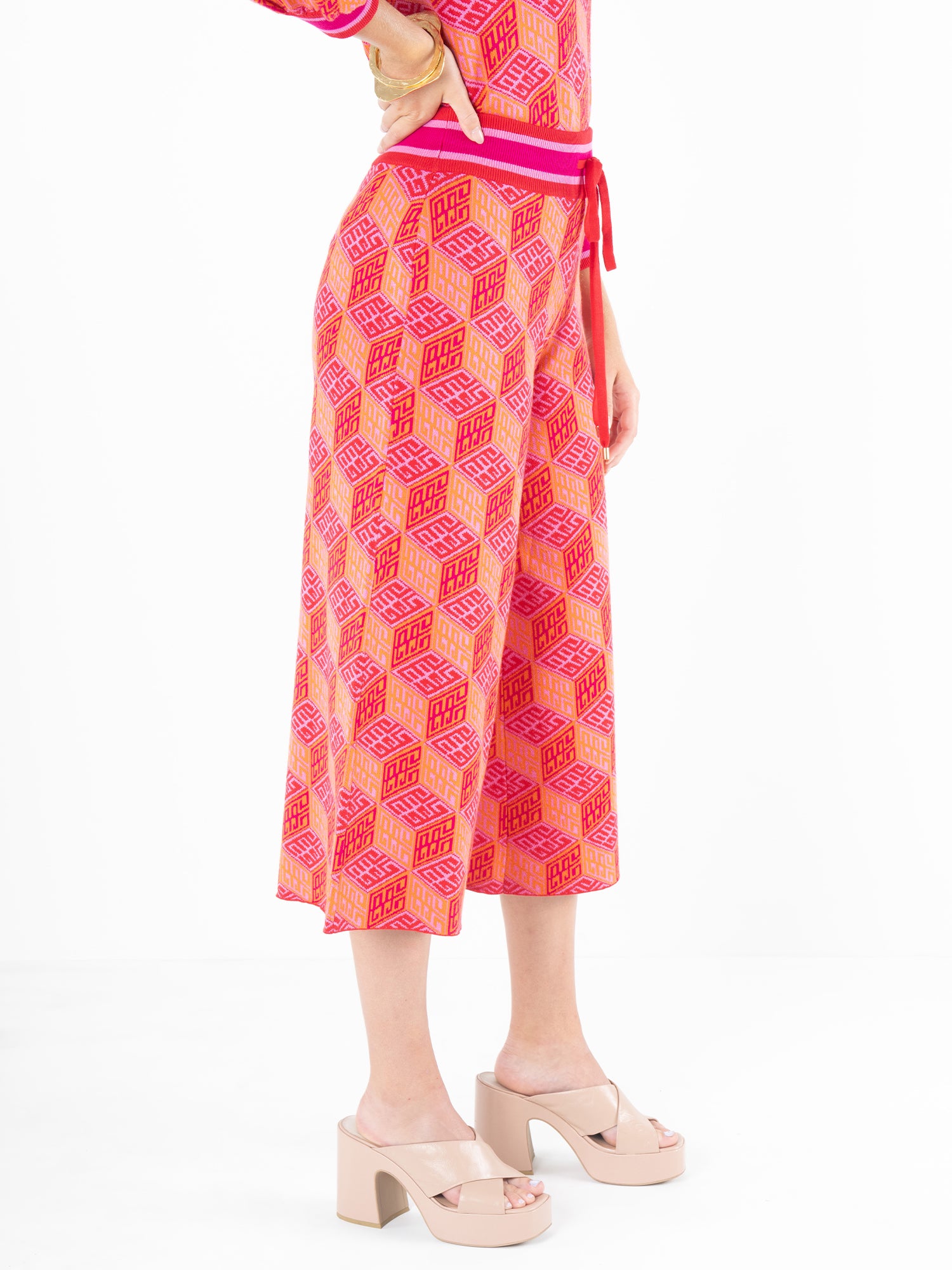 The individual is dressed in the Teddy Crop Trouser - Block Monogram, a pink geometric jumpsuit with cropped wide legs, styled elegantly with platform sandals against a white backdrop. This playful ensemble, made from breathable viscose knit, is accentuated by a striking red belt at the waist.