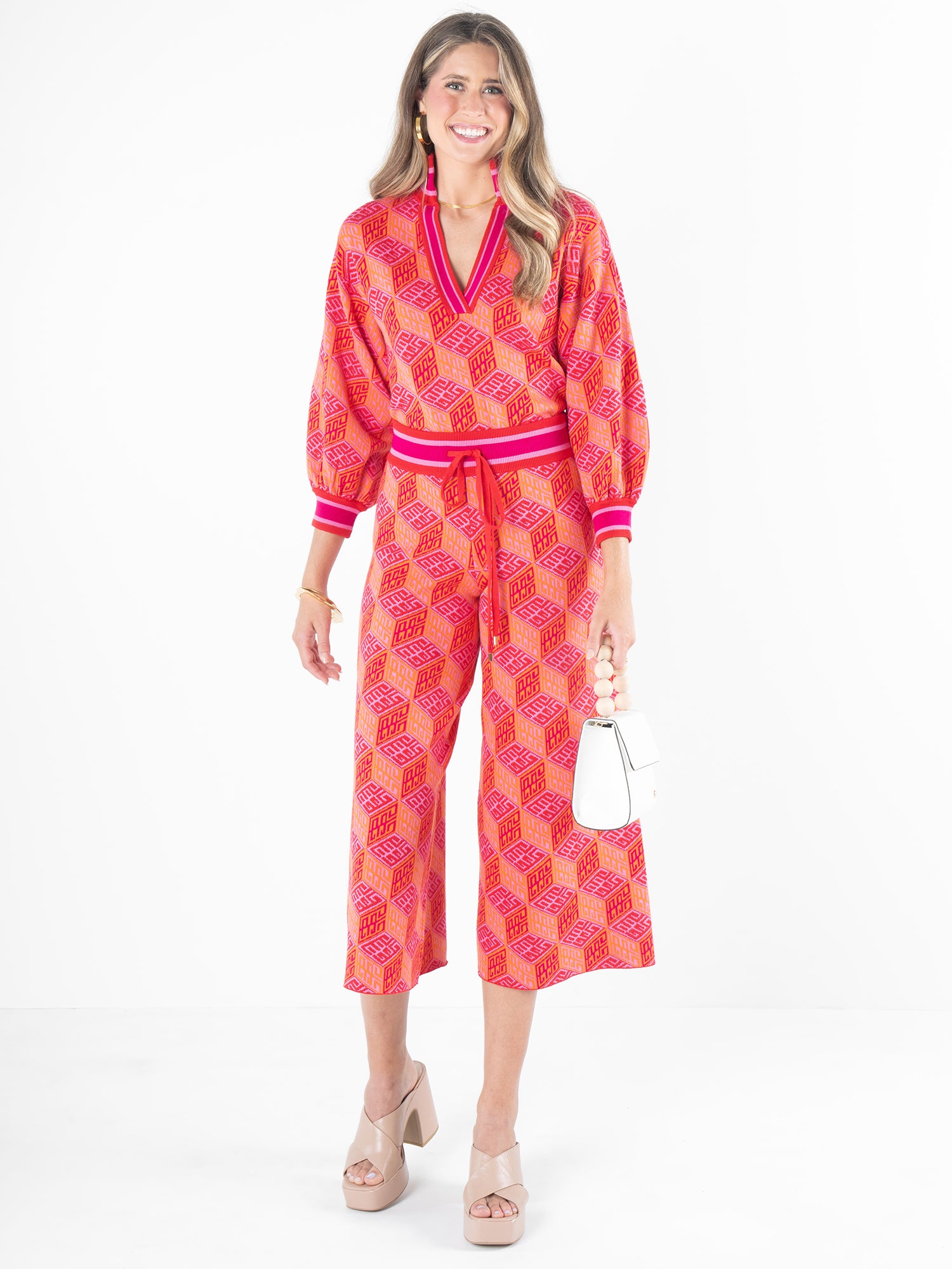 A woman beams, dressed in the Teddy Crop Trouser - Block Monogram, featuring vibrant pink and red geometric patterns with a tied waist and cropped wide legs. She holds a white handbag and wears beige high-heeled sandals, all set against a plain white background.